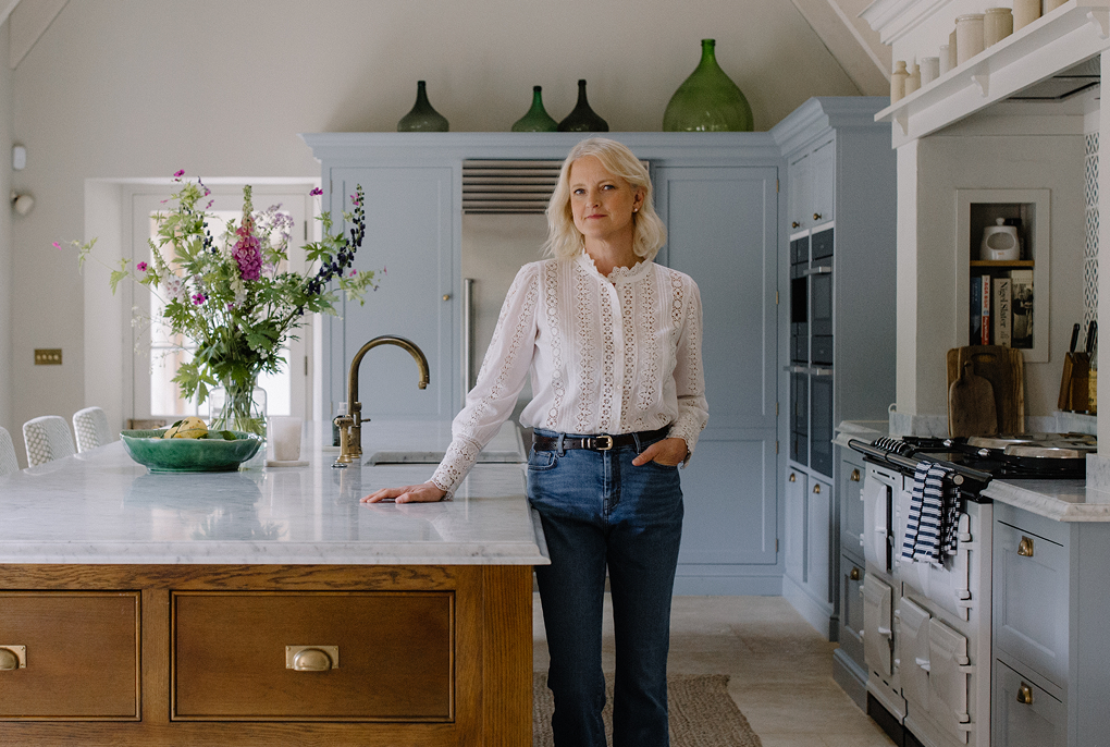 Designer Emma Simms-Hilditch stands in her kitchen with a hand on the Island, she is wearing a white top and blue jeans. 