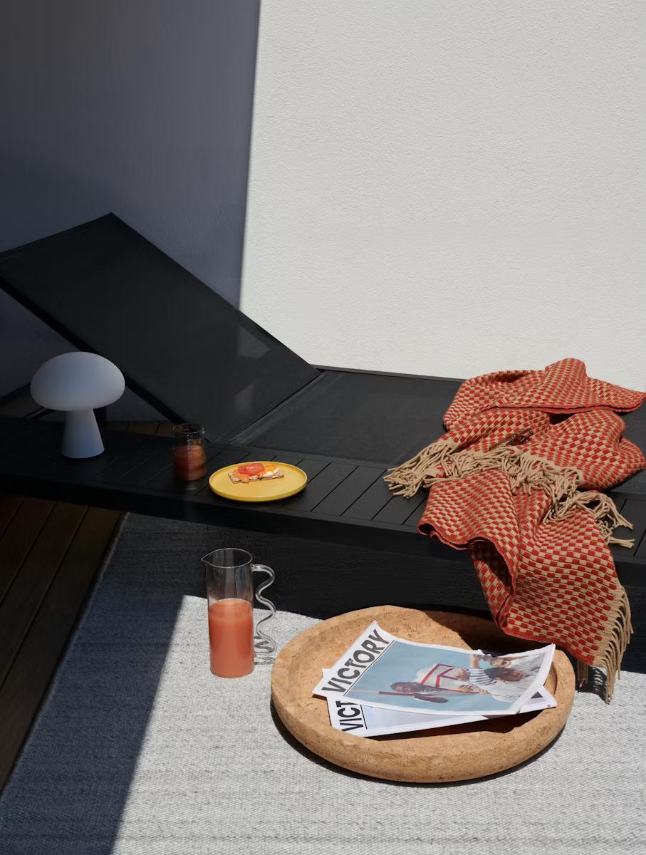  An outdoor lounge chair sits half in the shade. A red checked blanket is thrown over the foot of the chair, beside the chair is a pitcher of juice, and a large plate with magazines. 
