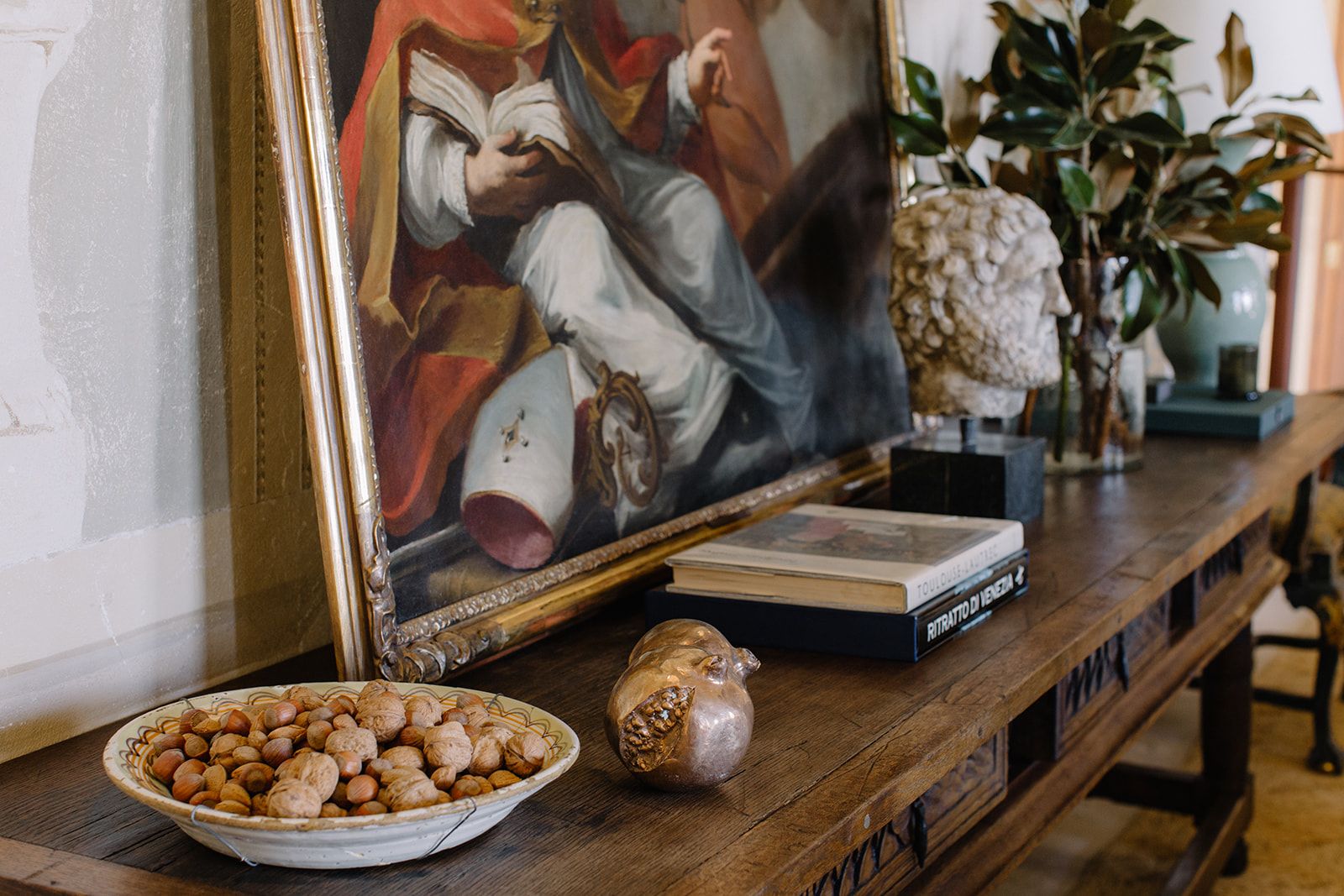 A wooden entryway table with a bowl of nuts, a brass pomegranate, a painting, a bust, and a few coffee table books.
