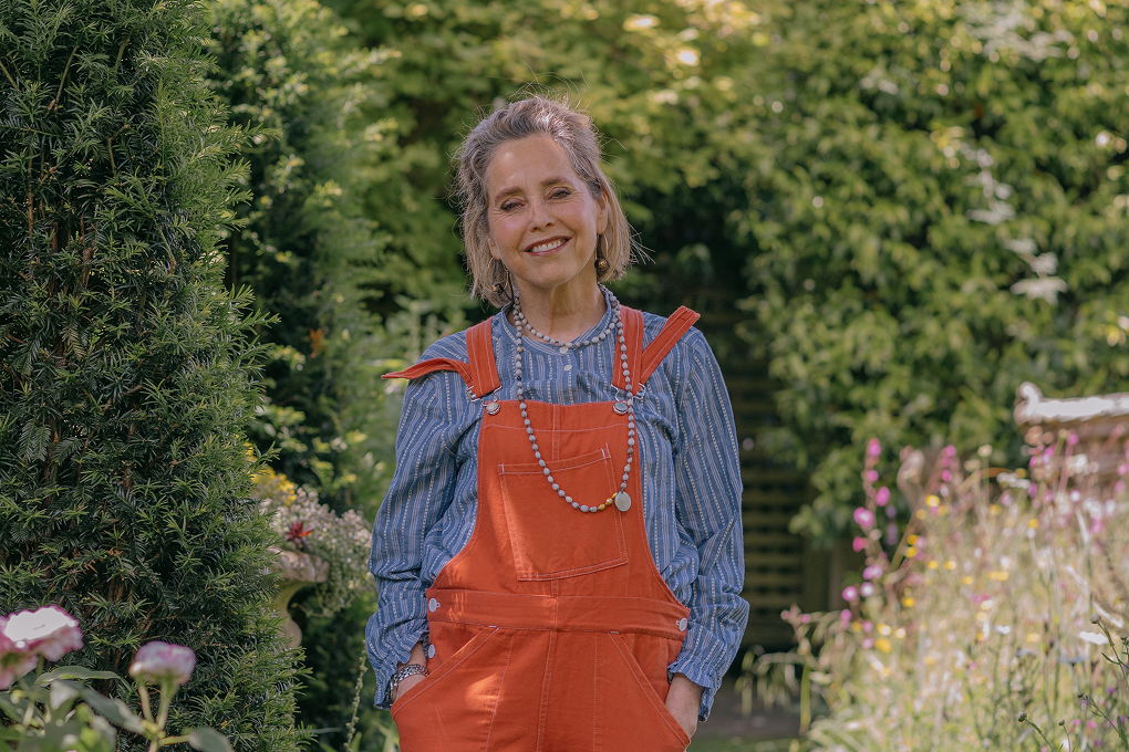 Garden designer Butter Wakefield stands in her garden. She is wearing orange overalls and a blue top with her hands in her pockets.