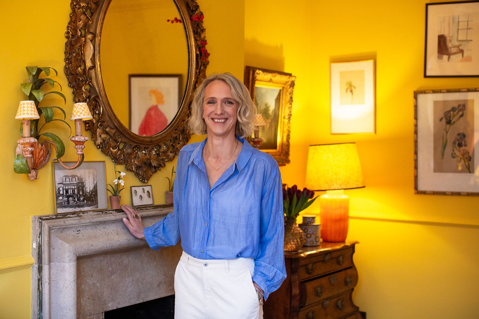 Lampshade designer Rosi de Ruig stands in front of the stone fireplace in her yellow living room. She wears a blue button down shirt and white trousers. 