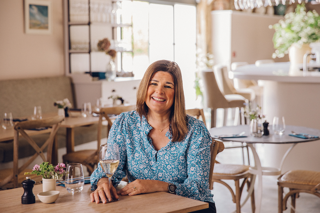Wine critic Helen McGinn sits at a restaurant table, she is holding a glass of champagne and smiling at the camera. 
