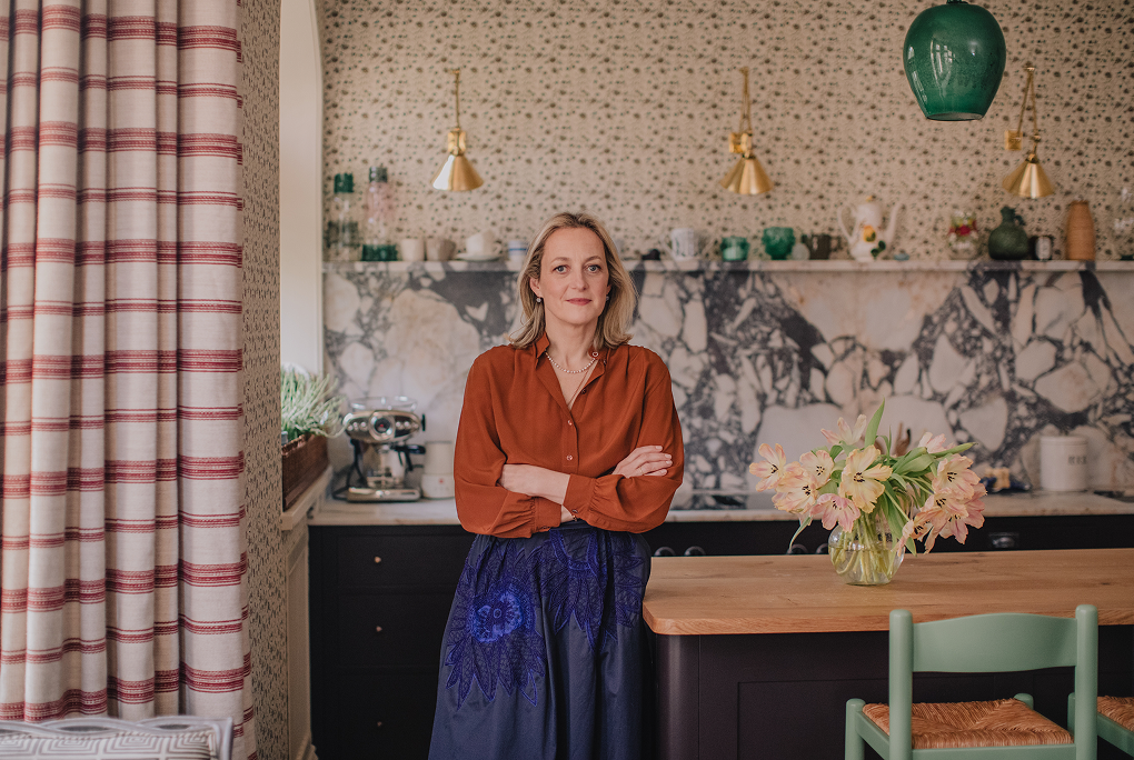 Designer Rita Konig stands in her kitchen with her arms crossed. She is wearing an orange top and a blue skirt. 