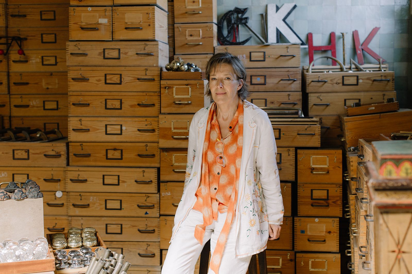 Designer Maria Speake sits on a stool in front of a wall stacked with salvaged wooden drawers. She is dressed in a bright orange top with a white jacket and trousers, looking directly at the camera. 