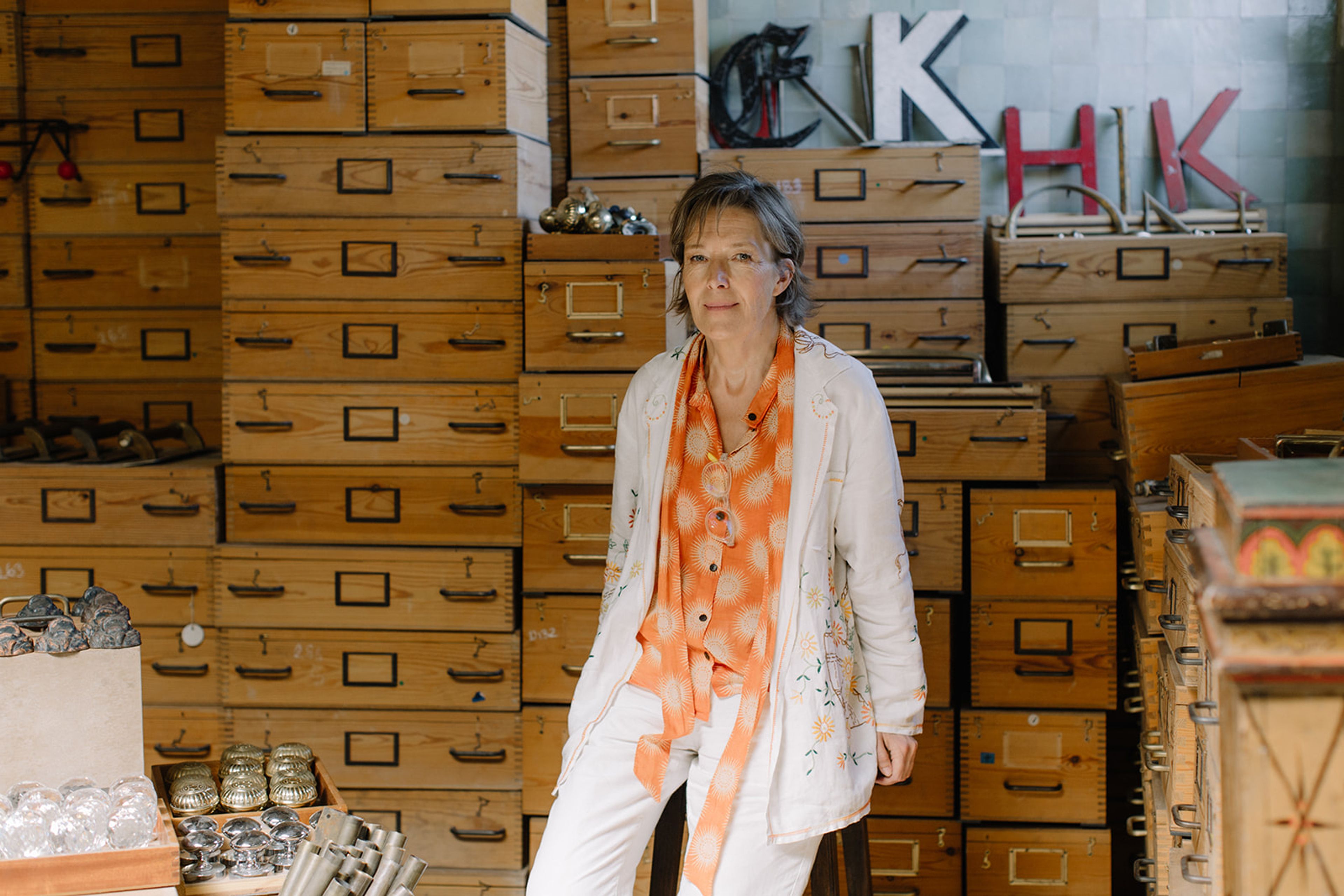 Designer Maria Speake sits on a stool in front of a wall stacked with salvaged wooden drawers. She is dressed in a bright orange top with a white jacket and trousers, looking directly at the camera. 