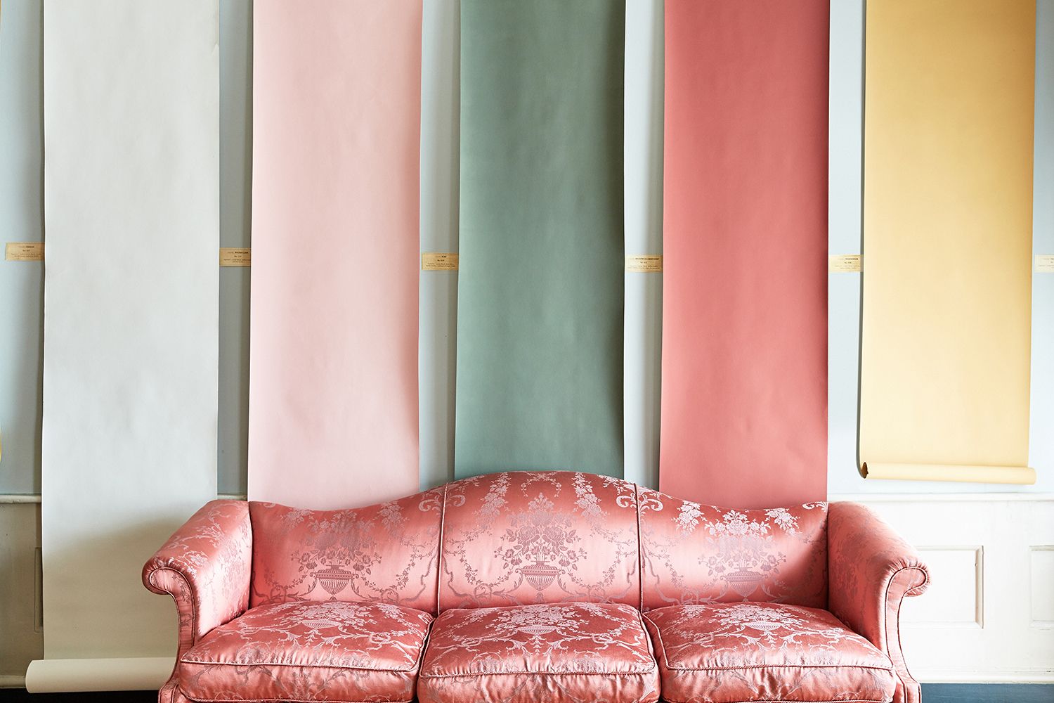 A pink satin sofa sits in front of five large sheets of Edward Bulmer paint swatches in white, pink, green, red, and yellow.