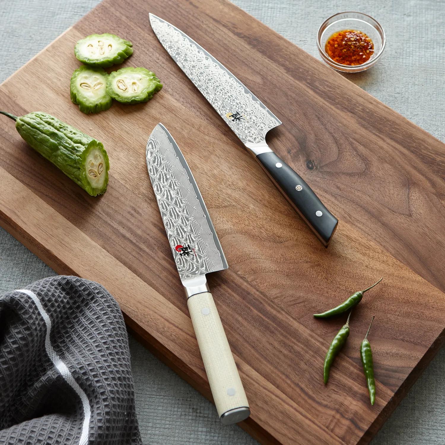 two Japanese-style chef's knives lay on a chopping board, in the top left is a cucumber that has been slices, in the bottom right are some green chillis. 