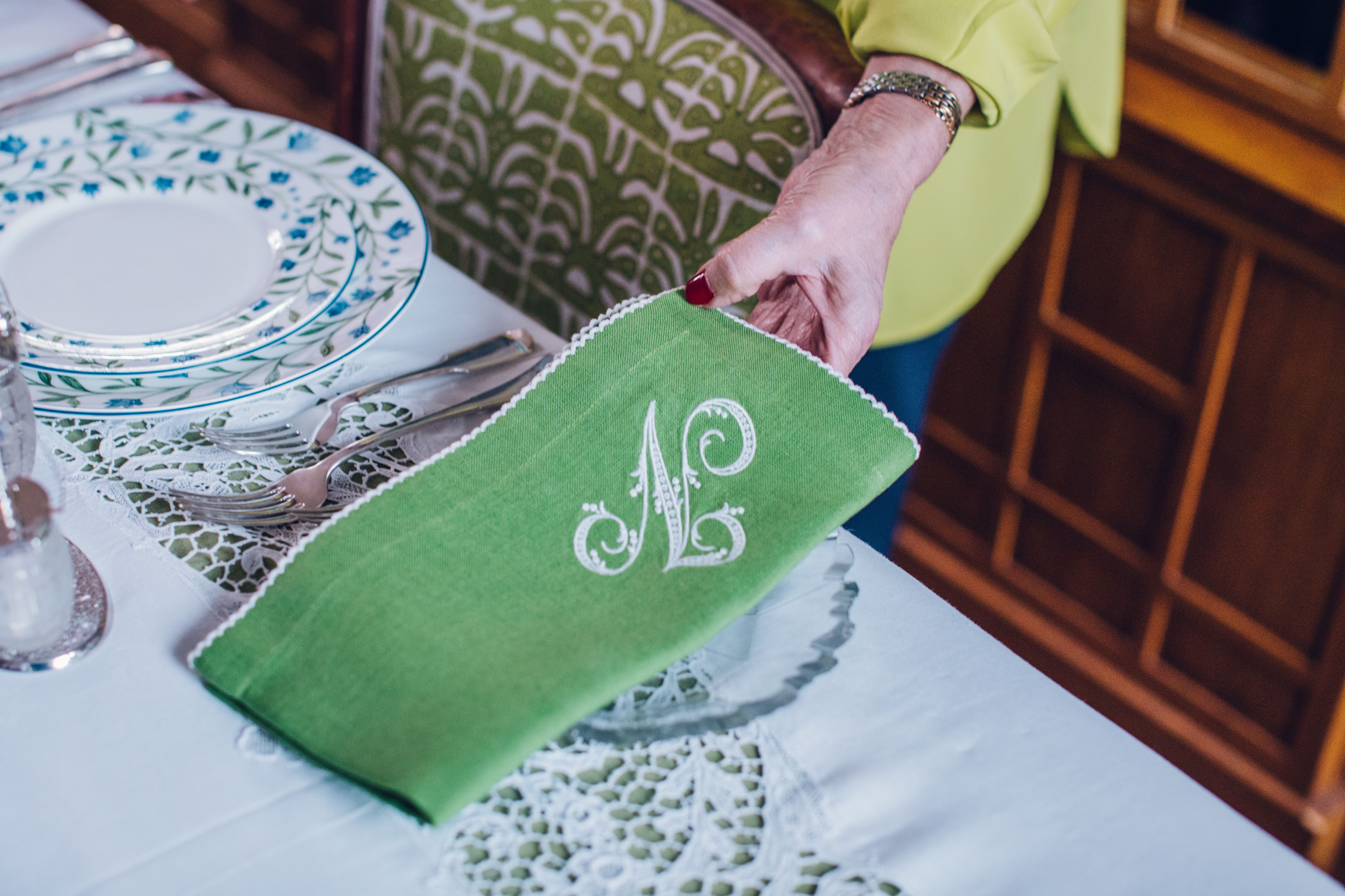 A hand lays a green napkin embroidered with a white N on a beautifully set table. 