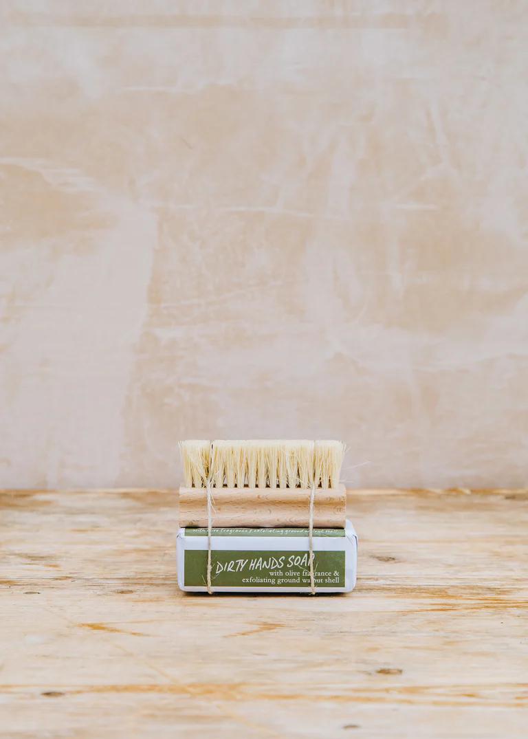 A product image of a natural wood and bristle nail brush artfully tied to packaged bar soap.