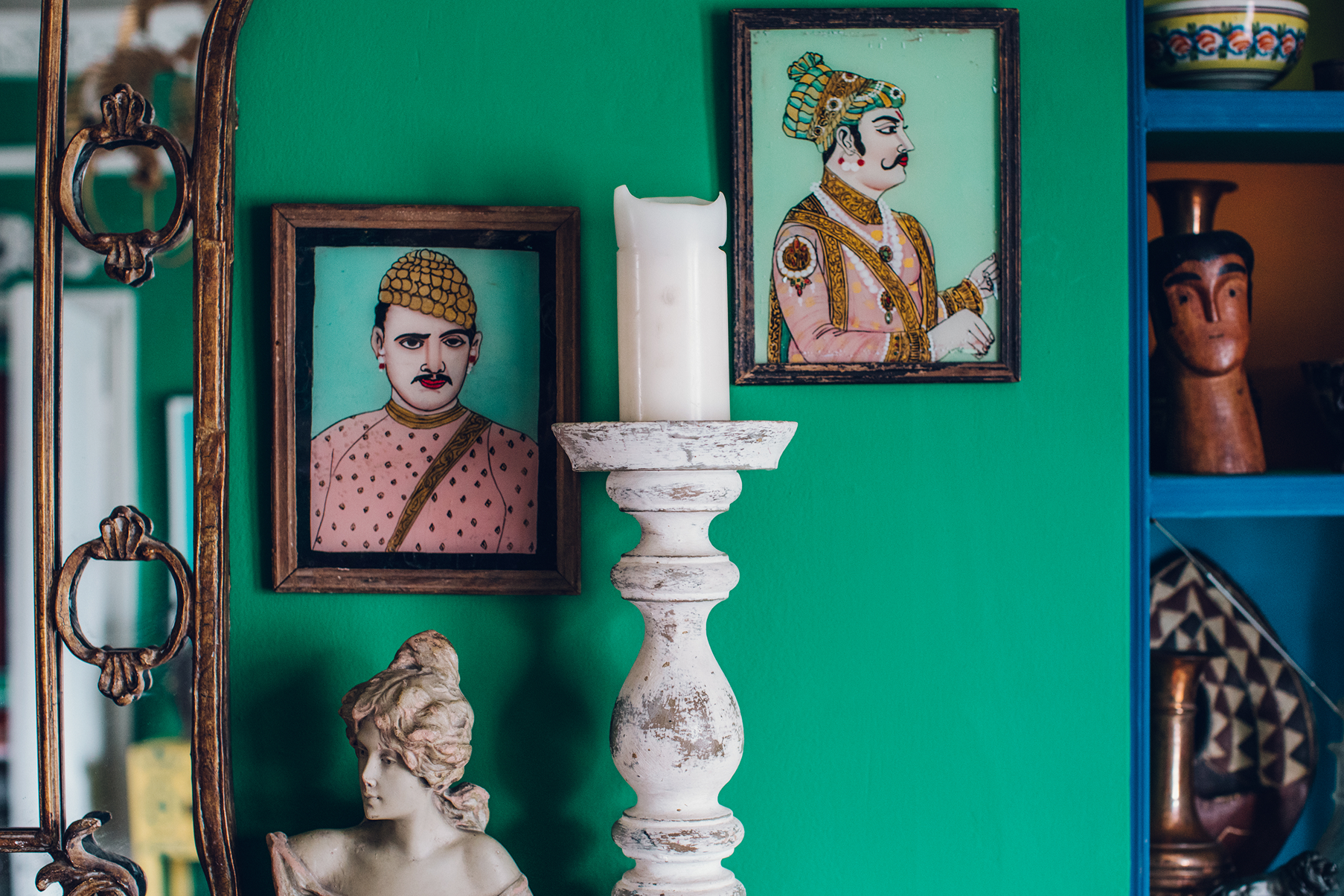 A close up of Annie Sloan's living room. The wall is painted bright green. In the centre is a tall wooden candlestick painted white, on either side are vintage paintings. 
