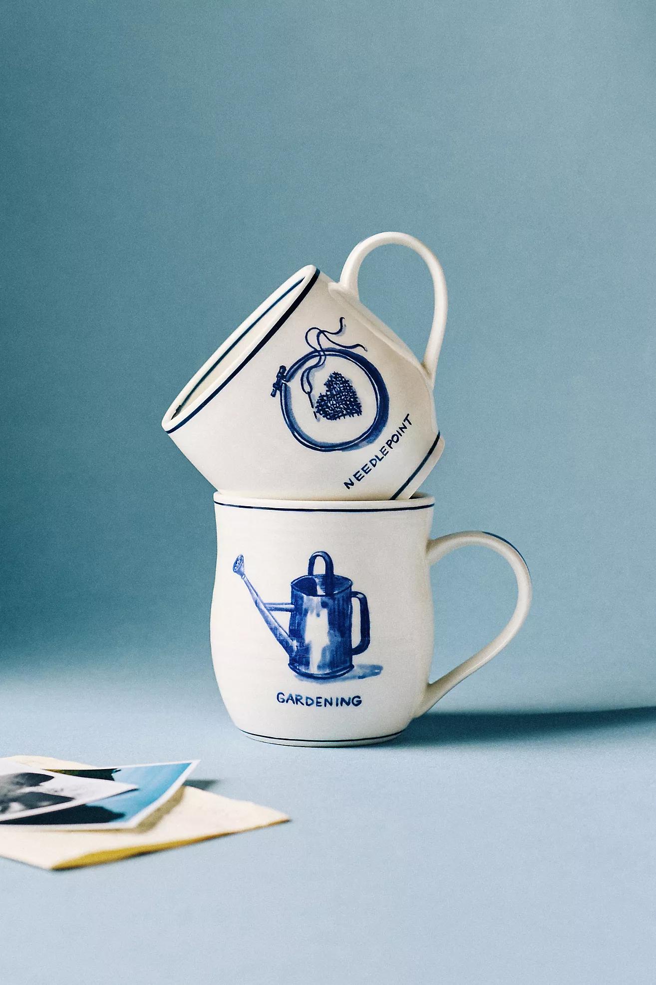 Two stoneware mugs from Anthropolige. One mug has a picture of a needlepoint heart with the worlds 'Needlepoint' and the other has a picture of a watering can with the words 'gardening' 