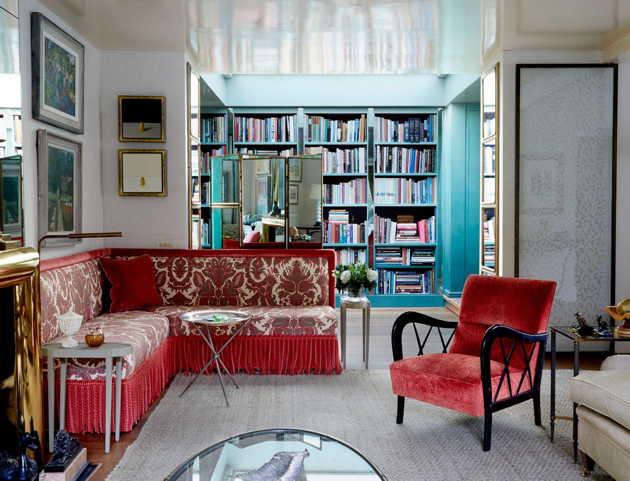 A view of Nina Campbell's living room and entryway. A red tufted L-shaped sofa sits in front of a large turquoise bookshelf.