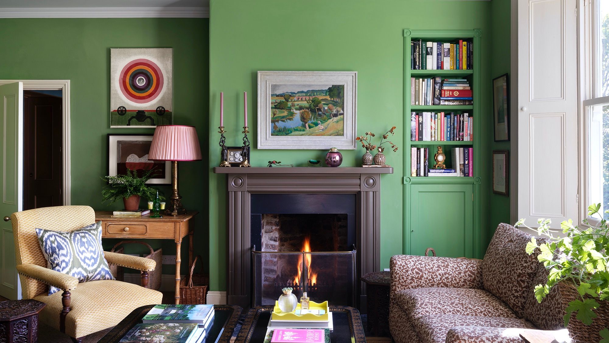 Rita’s living room painted in Edward Bulmer’s Invisible Green. In the centre of the room is a burning fireplace, to the right is a bookshelf, and in the foreground are a sofa and chair.
