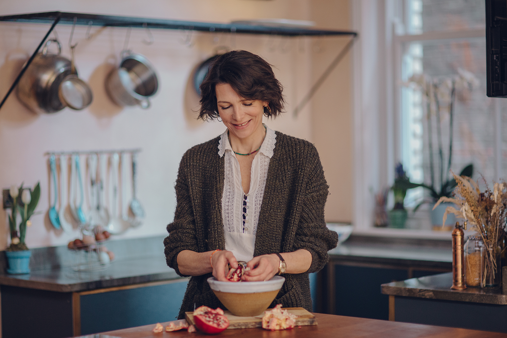 Chef Thomasina Miers stands over a large bowl. She is holding a pomegranate and removing the seeds. 