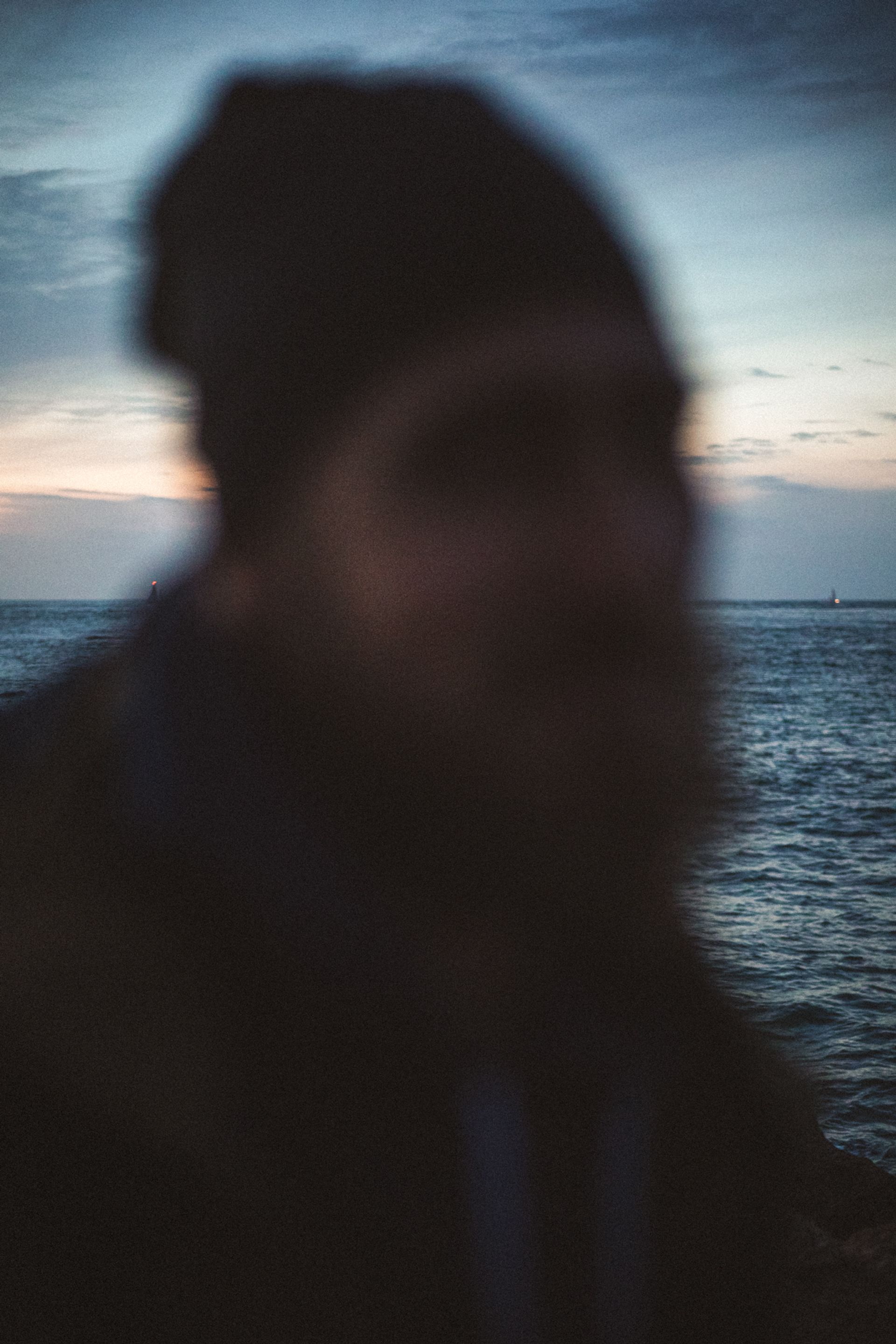 A blurred figure of a person with a beanie stands near a body of water at dusk, with a pastel sky in the background.