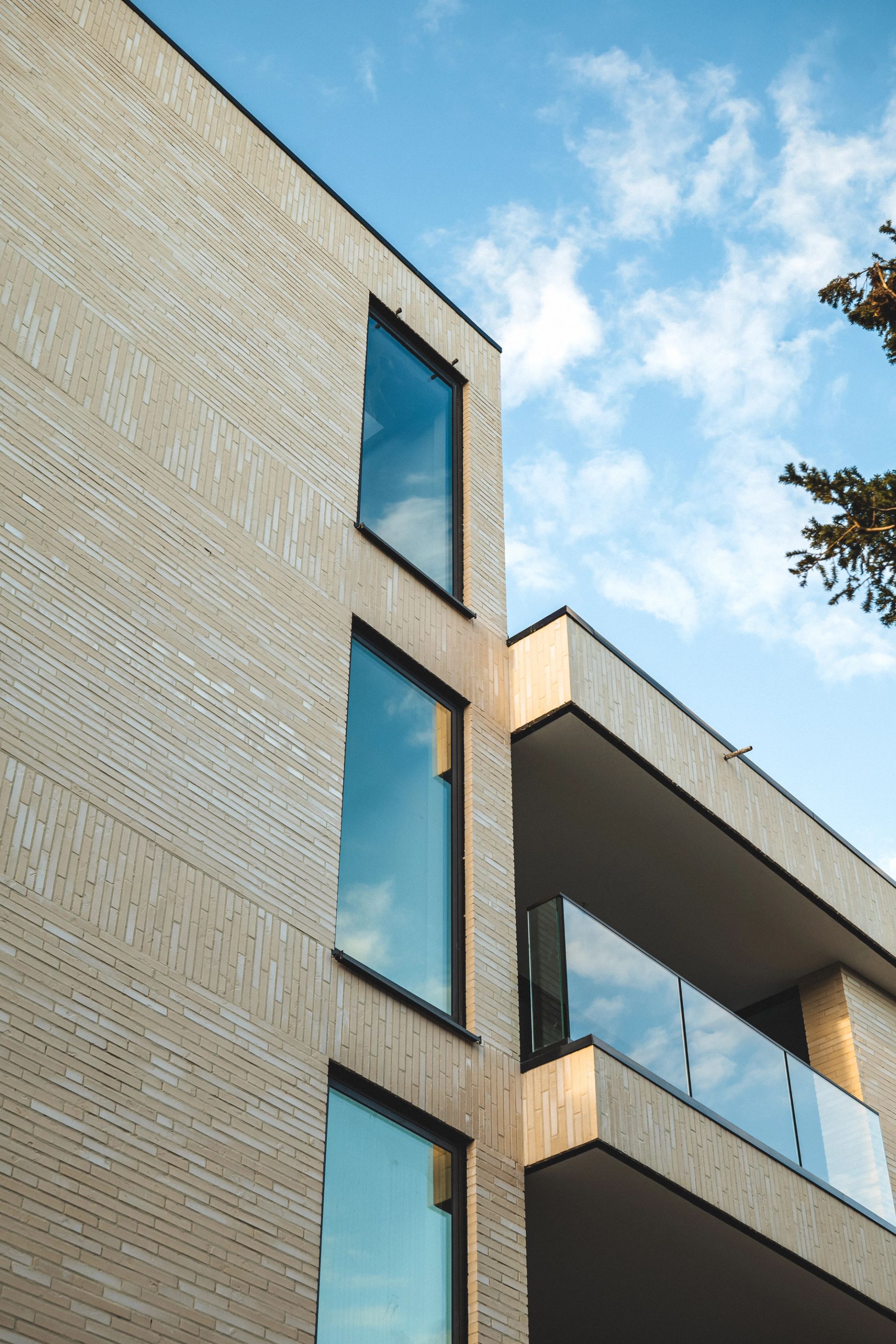 Exterior facade of a modern residential building with light-coloured brick cladding, floor-to-ceiling windows and a cantilevered balcony photographed against a blue sky, Belgium.