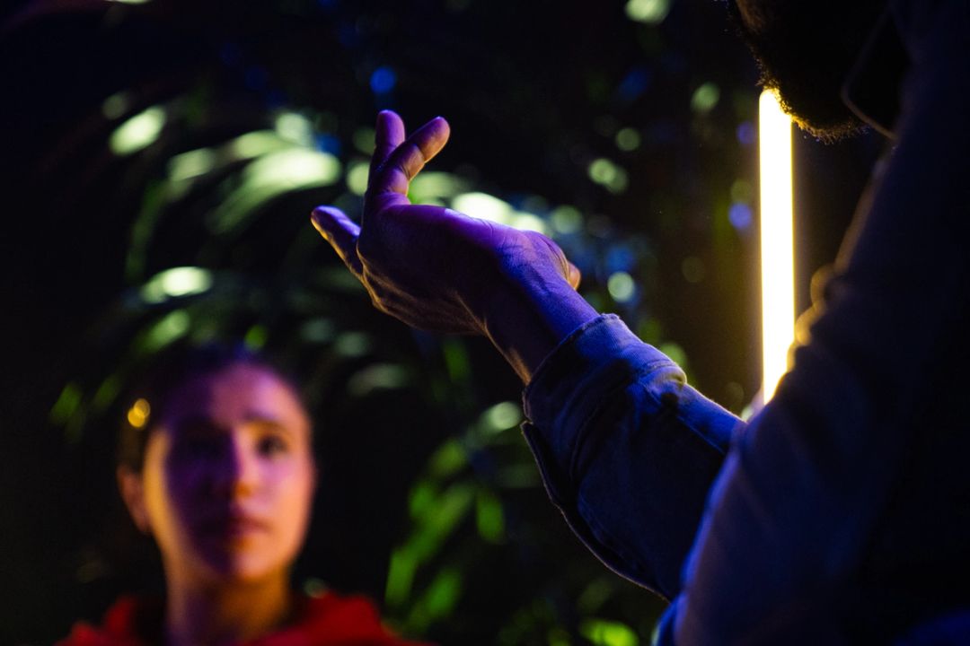 Gros plan d'une main ouverte tournee vers le haut, poignet en jean, visage d'une femme flou derriere pres d'un tube lumineux chaud.
