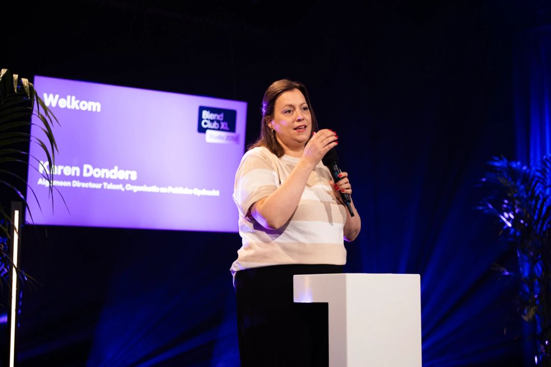 Karen Donders, en haut rayé, parle au micro devant un pupitre; la diapo de bienvenue et le logo Blend Club XL brillent derrière.