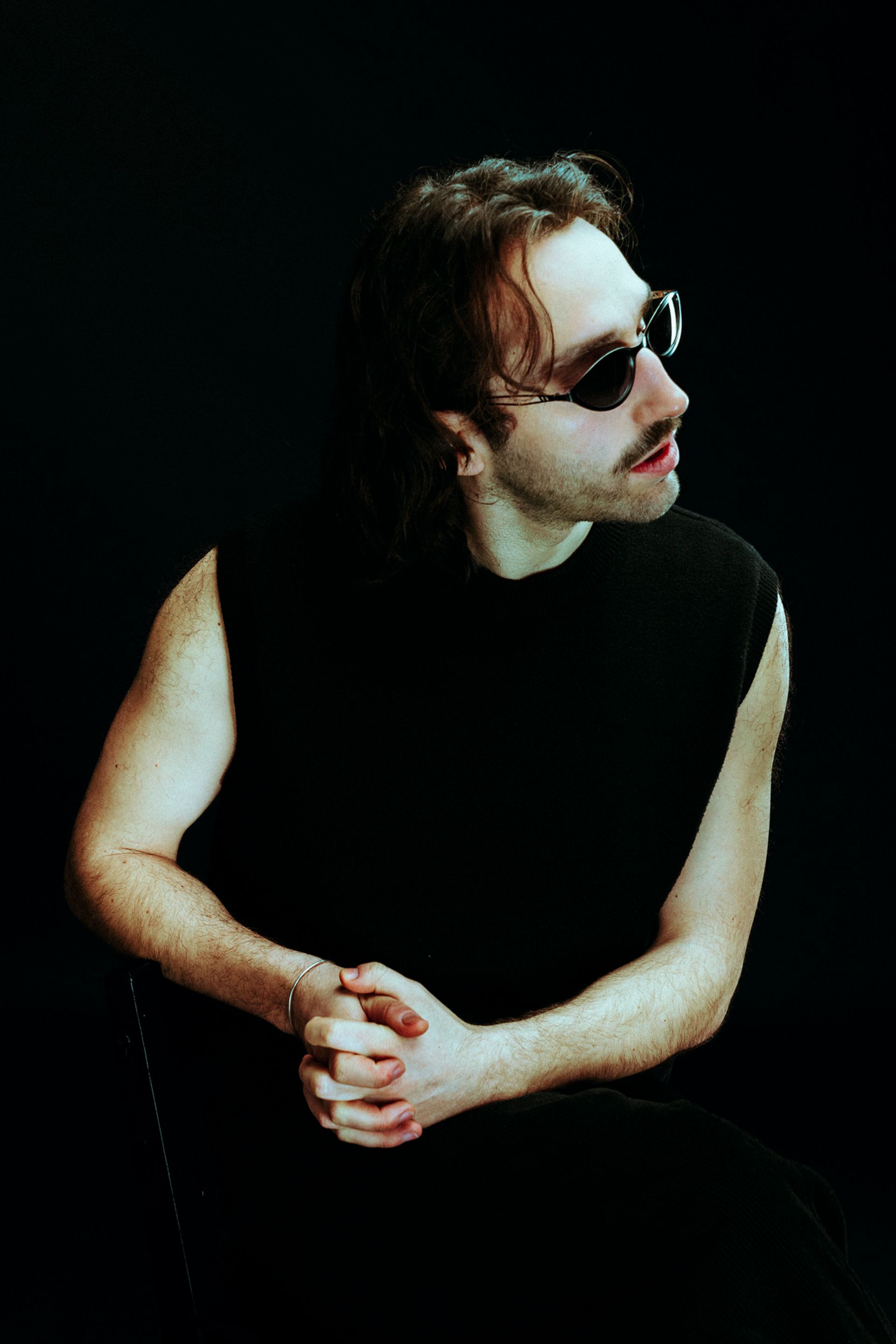 Un homme assis, portant un gilet noir et des lunettes de soleil, se tourne légèrement vers la gauche.