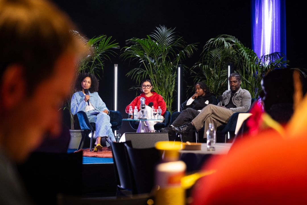 Panel de quatre assis sur scene parmi les palmiers, la moderatrice en jean avec un micro, silhouettes du public au premier plan.