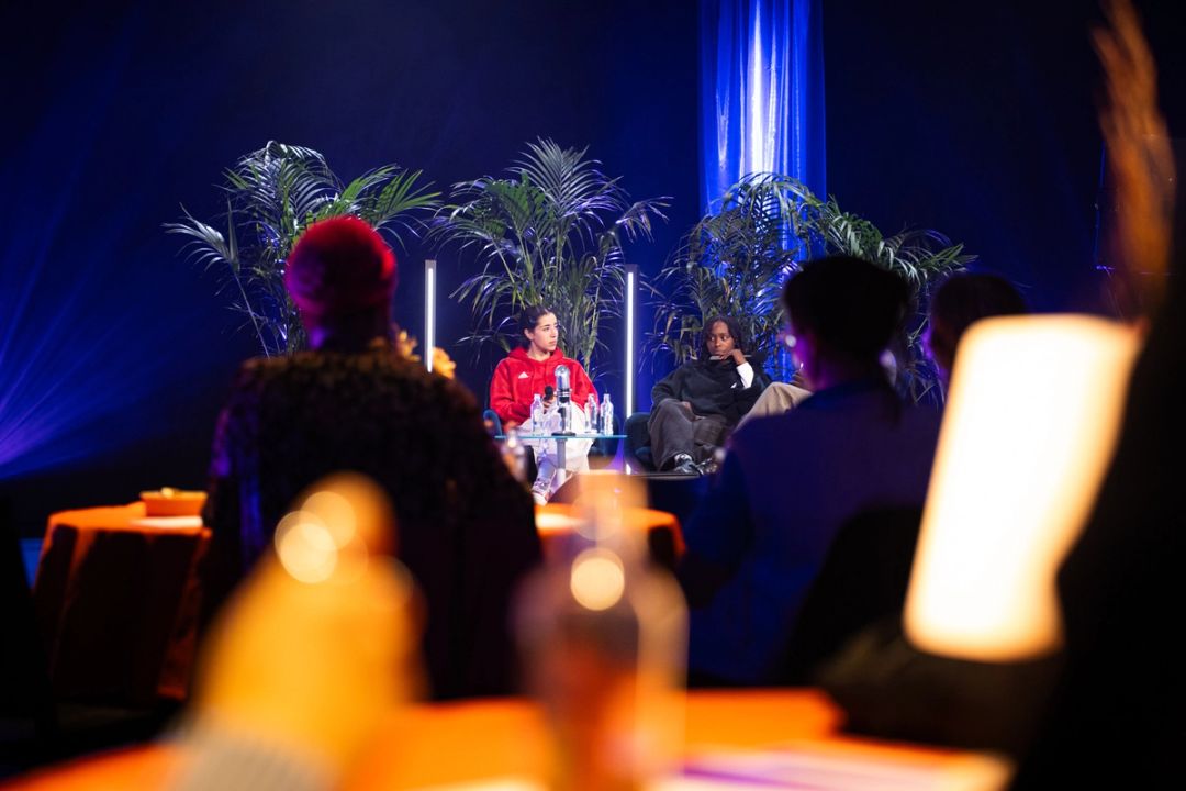 Panel de deux femmes vu derriere le public, l'une en sweat rouge et l'autre en noir, lampes chaudes floues au premier plan.
