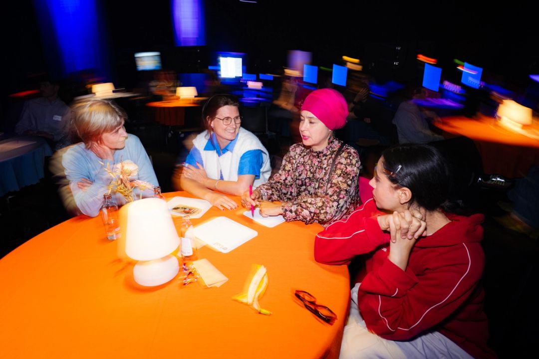 Quatre femmes discutent a une table ronde orange avec lampe, peau de banane et papiers, la salle floue en mouvement derriere elles.