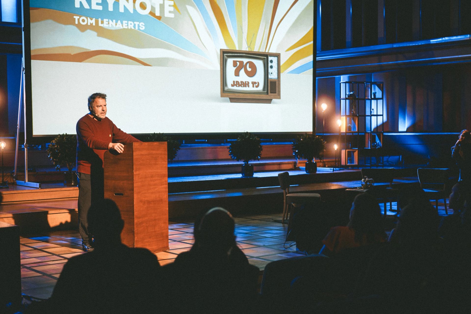 Tom Lenaerts speaks at a lectern during his keynote at the 70 jaar TV event, branding on screen behind him.
