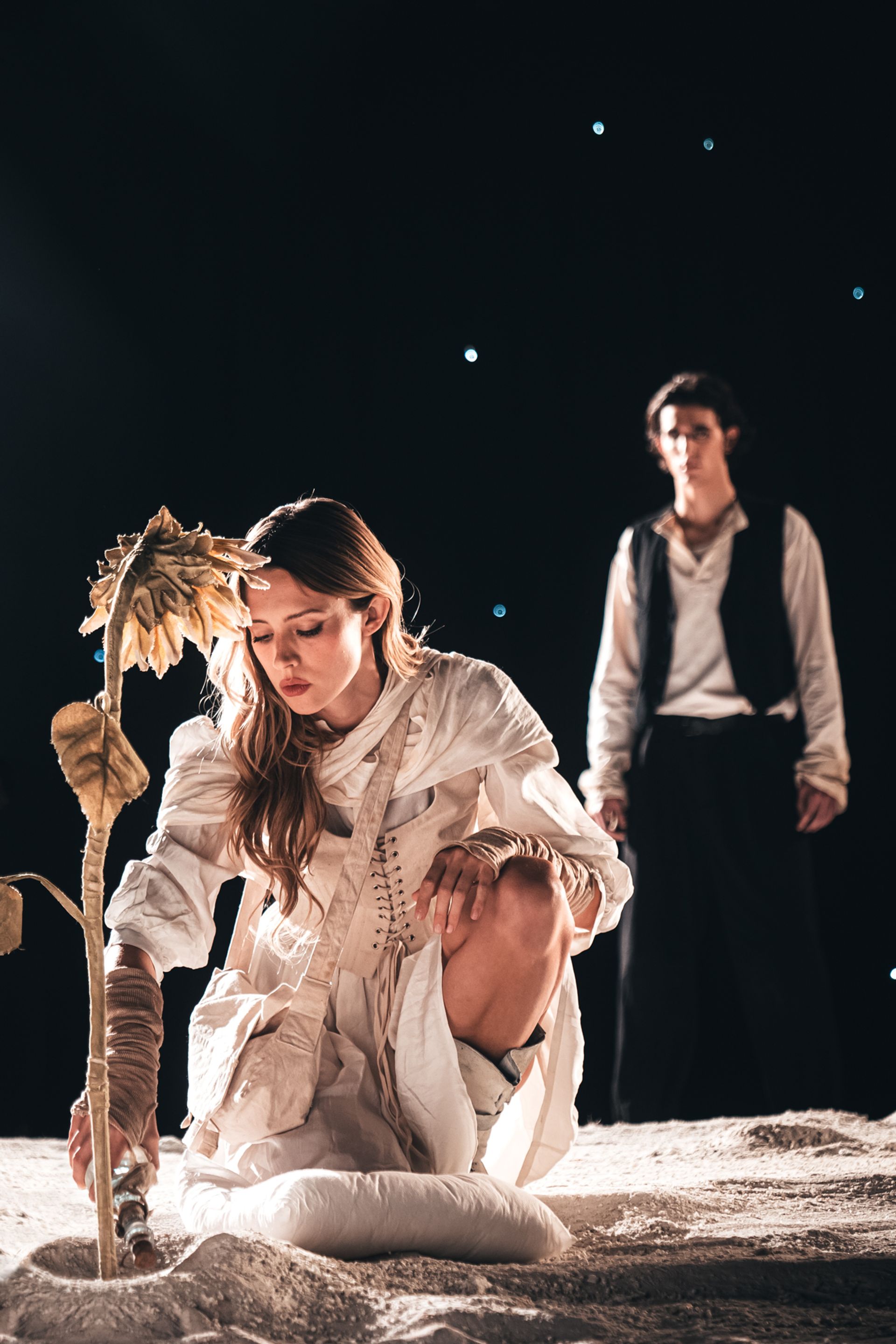 Angèle agenouillée sur une surface lunaire, penchée vers un tournesol fané, avec Tamino debout en arrière-plan sous un ciel sombre parsemé d'étoiles.