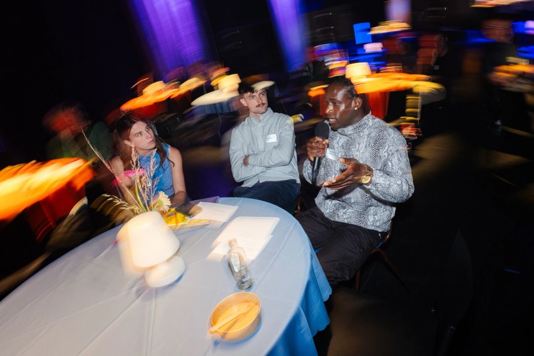 Table grand angle avec une femme en bleu, un homme en chemise rayee et un homme en chemise a motifs parlant au micro, nappe bleue et lampe.