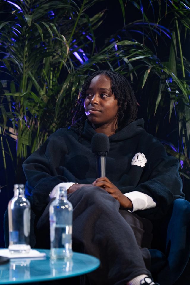 Une femme aux locks en sweat noir est assise au panel, bouteilles d'eau sur table en verre au premier plan, palmiers et lumiere bleue.