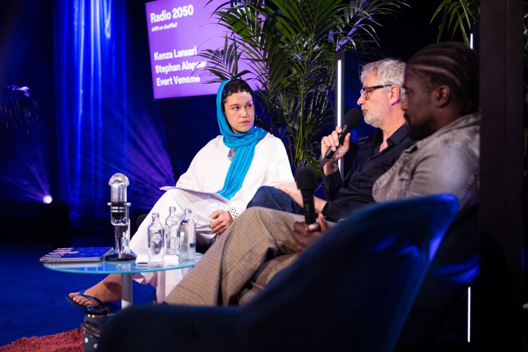 Discussion en panel; un homme grisonnant a lunettes parle au micro pres d'une femme au foulard bleu. L'ecran affiche Radio 2050.