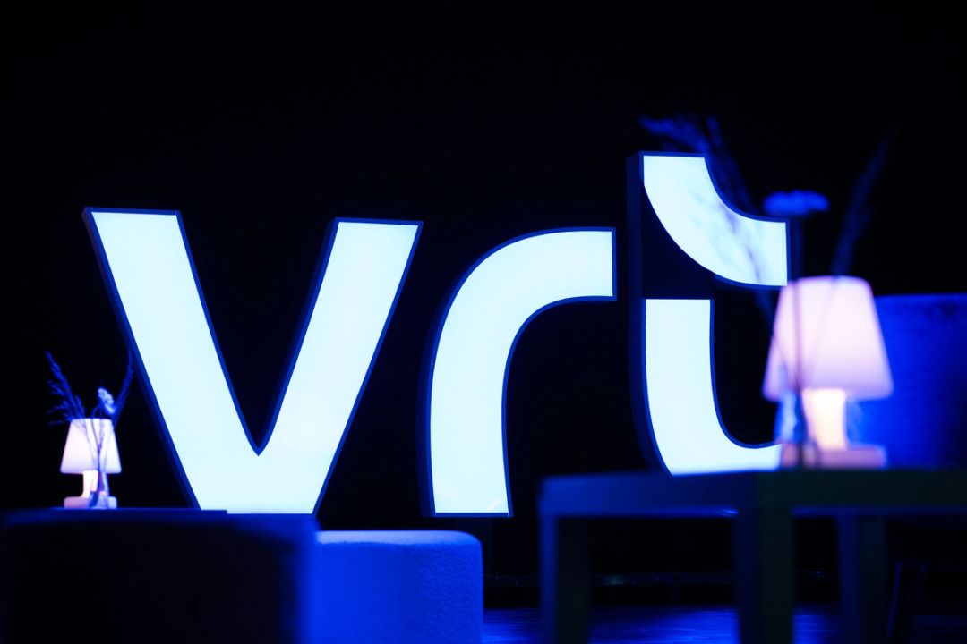 Les grandes lettres lumineuses blanches du logo VRT brillent sur la scène sombre de Blend Club XL, avec petites lampes et lumière bleue.