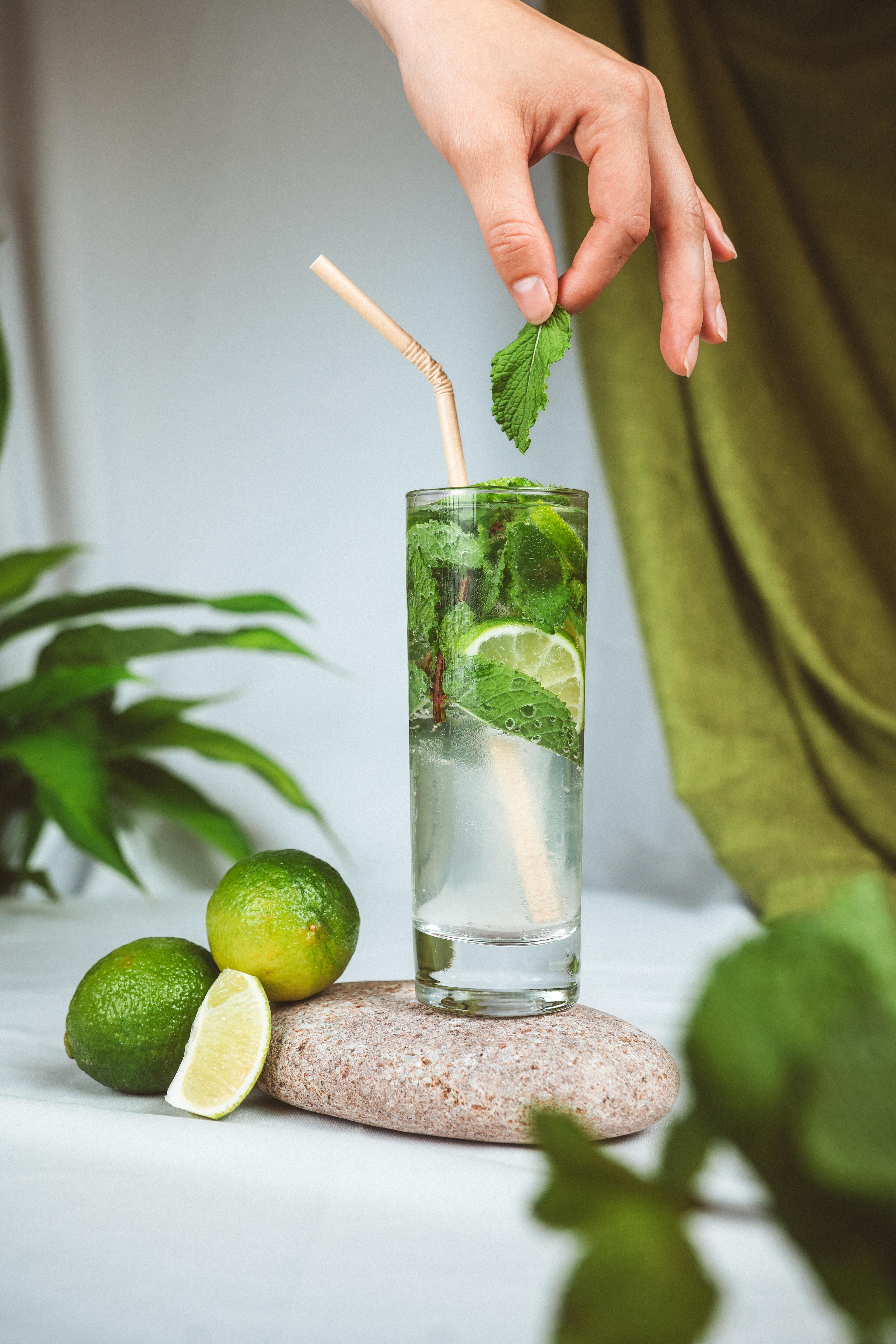 A transparent cocktail filled with pieces of lime and mint in a tall glass with a paper straw. The glass sits on a rock, surrounded by whole limes, against a white and green background.