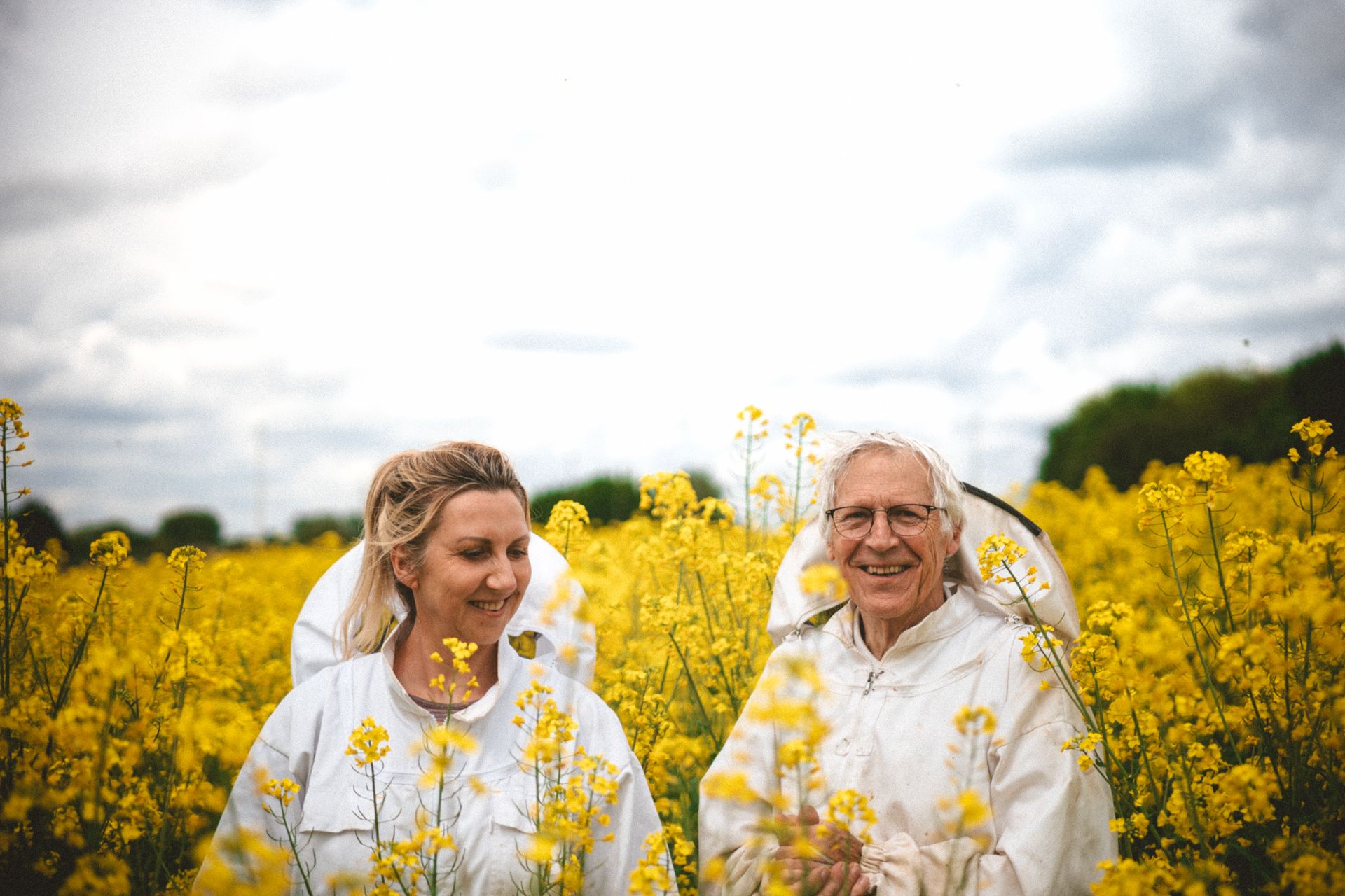 Deux apiculteurs souriants posent devant un champ de colza jaune. Un ciel nuageux en arrière-plan.