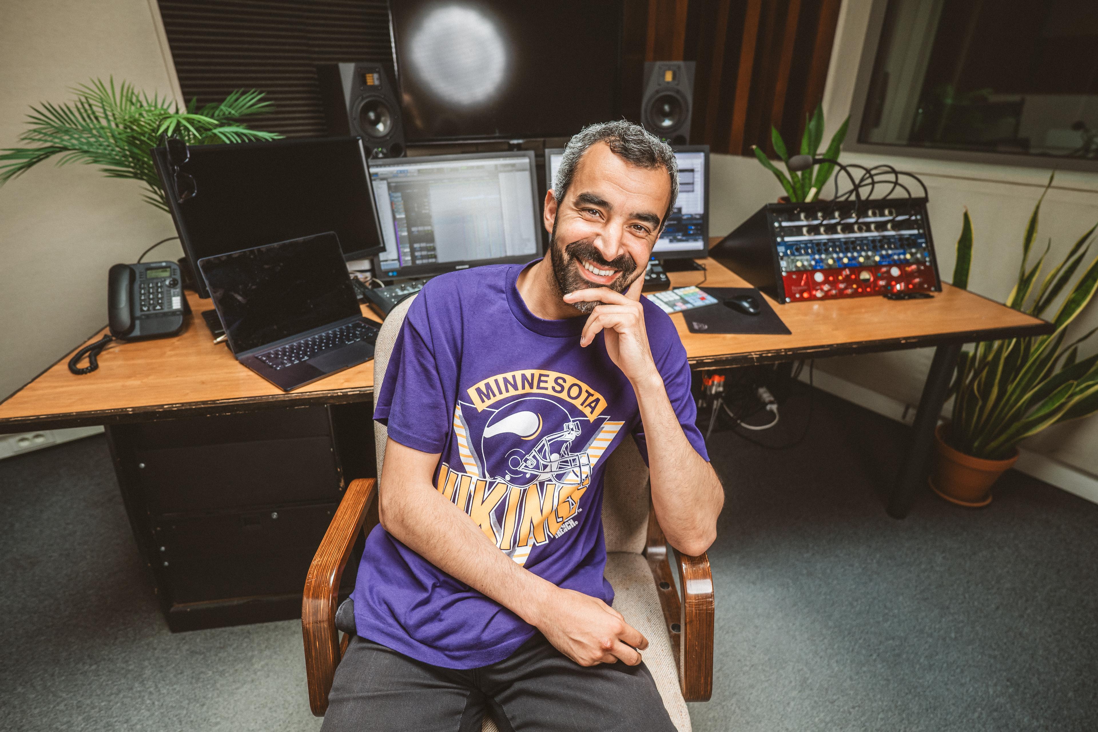 a middle aged man with a purple t-shirt sitting in a chair in front of his desk while leaning on his left arm