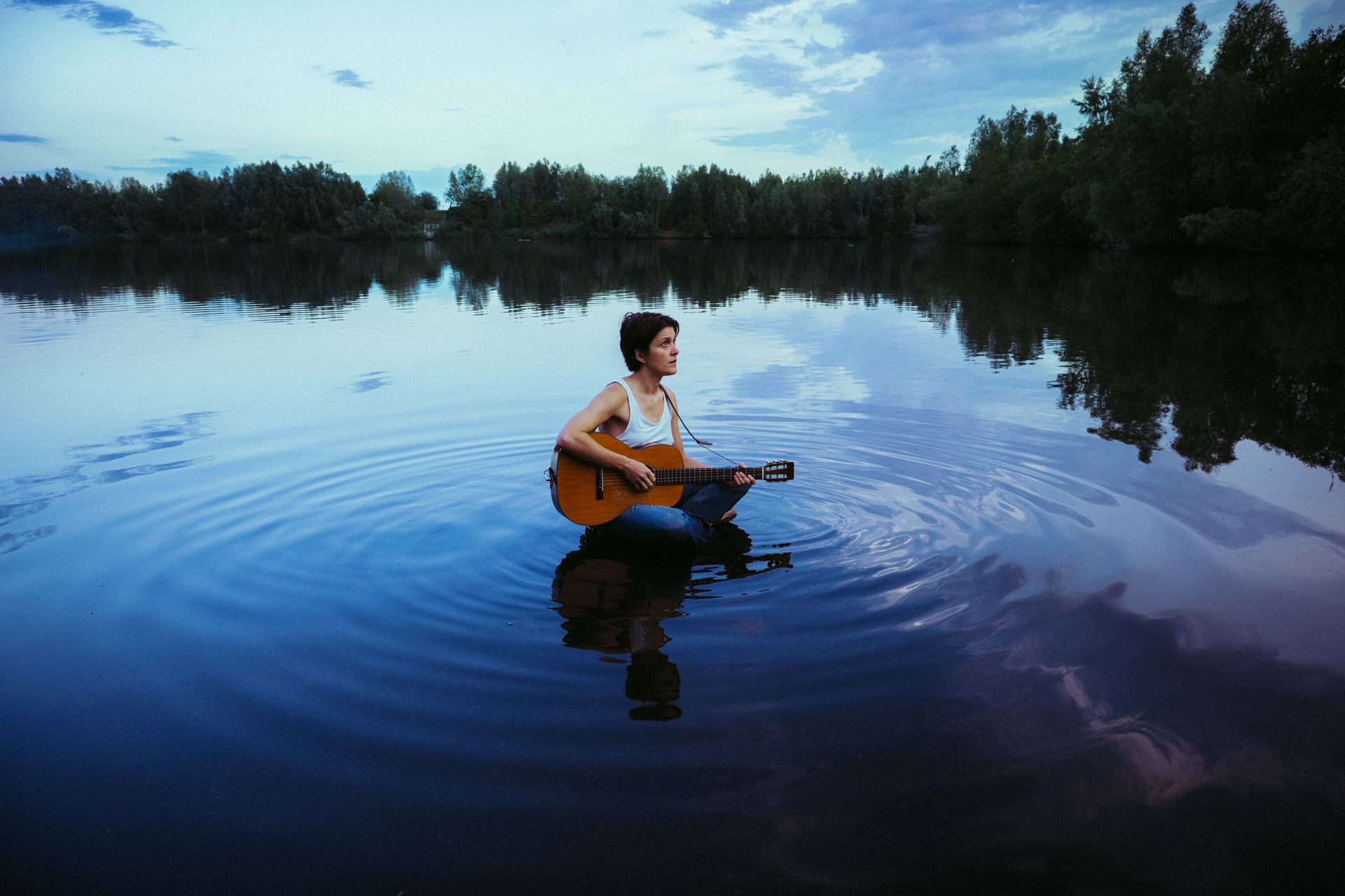 Une personne joue de la guitare dans un plan d'eau paisible, créant des ondulations sur la surface réfléchissante.