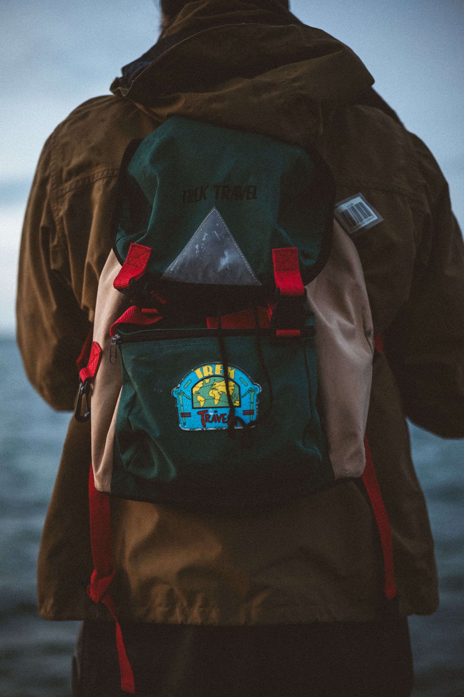 A person with a green and beige backpack stands facing the water, wearing a brown jacket with a hood.