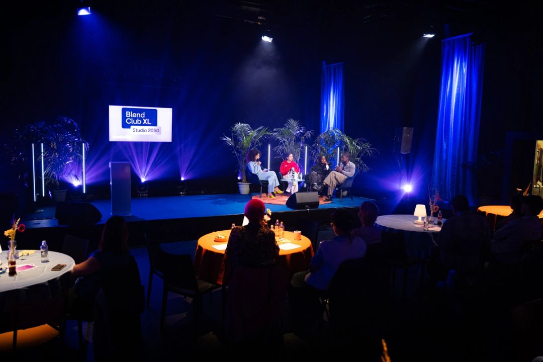 Vue large de la salle: un panel de quatre sur scene eclairee, l'ecran Blend Club XL Studio 2050, public aux tables rondes.