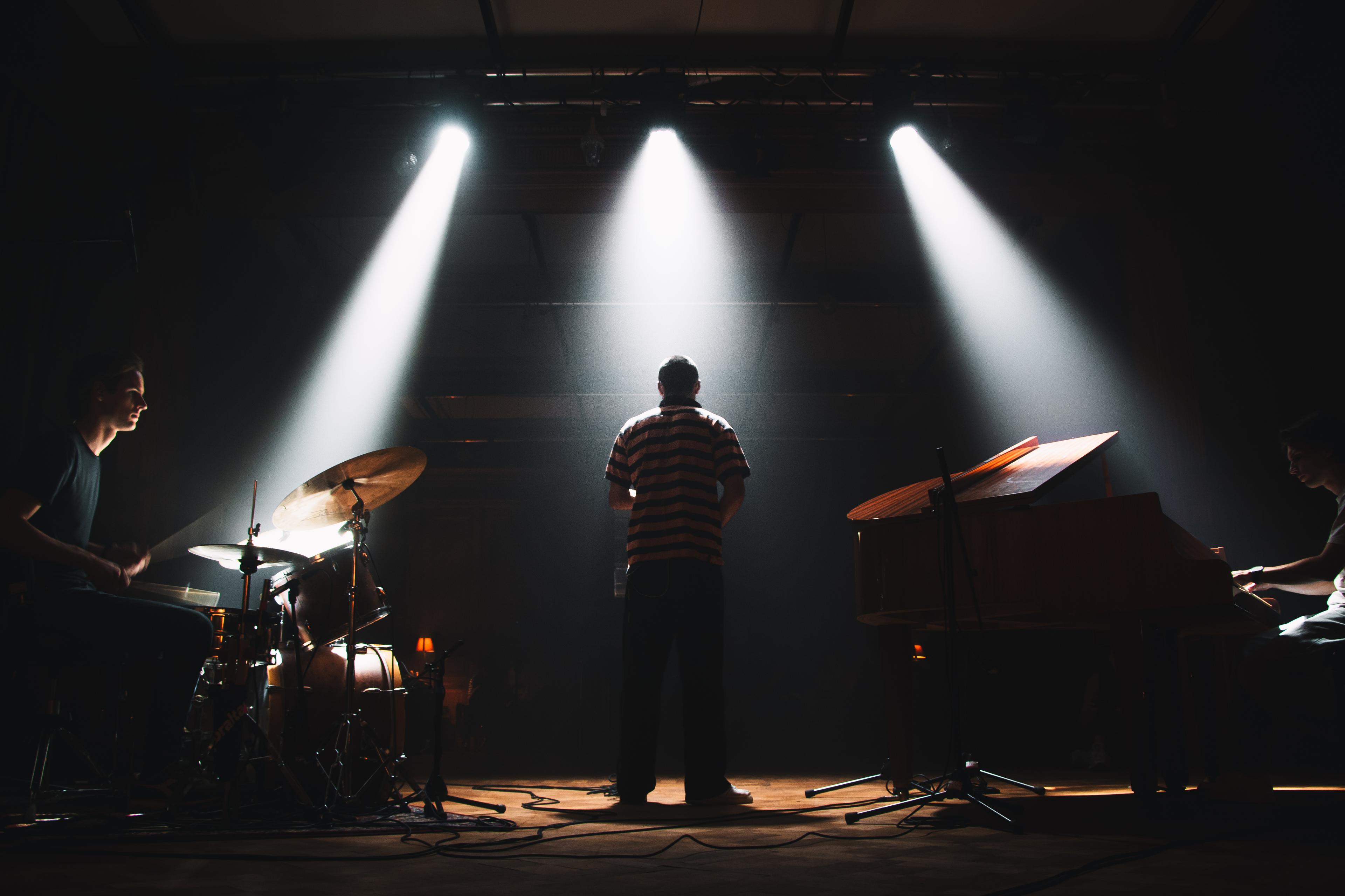 Silhouette of 3 guys standing in 3 different spotlights. One on the drums, one holding a saxophone and one on a big piano.