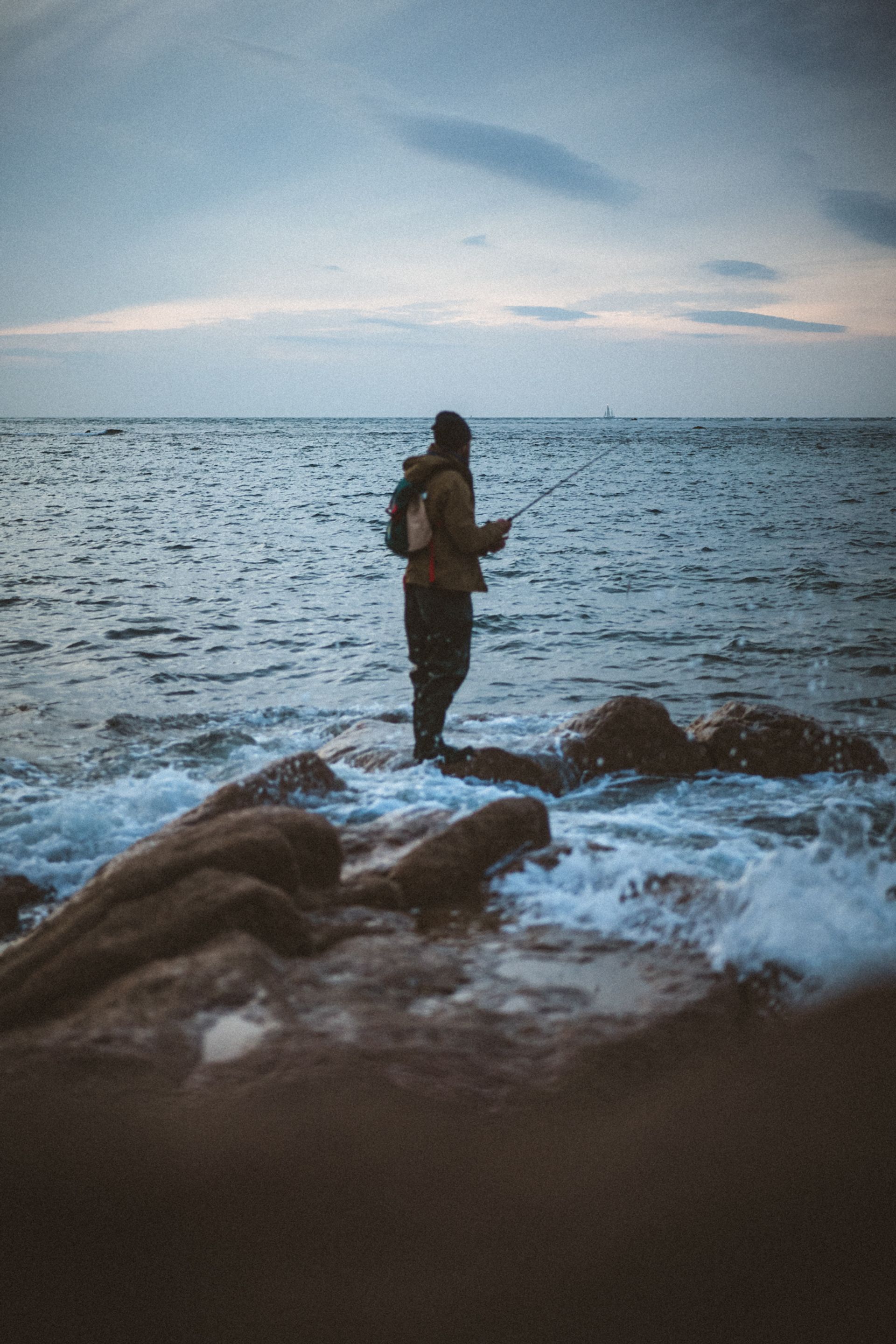 A person stands on rocky shore, fishing in the ocean with waves crashing around them under a cloudy sky.