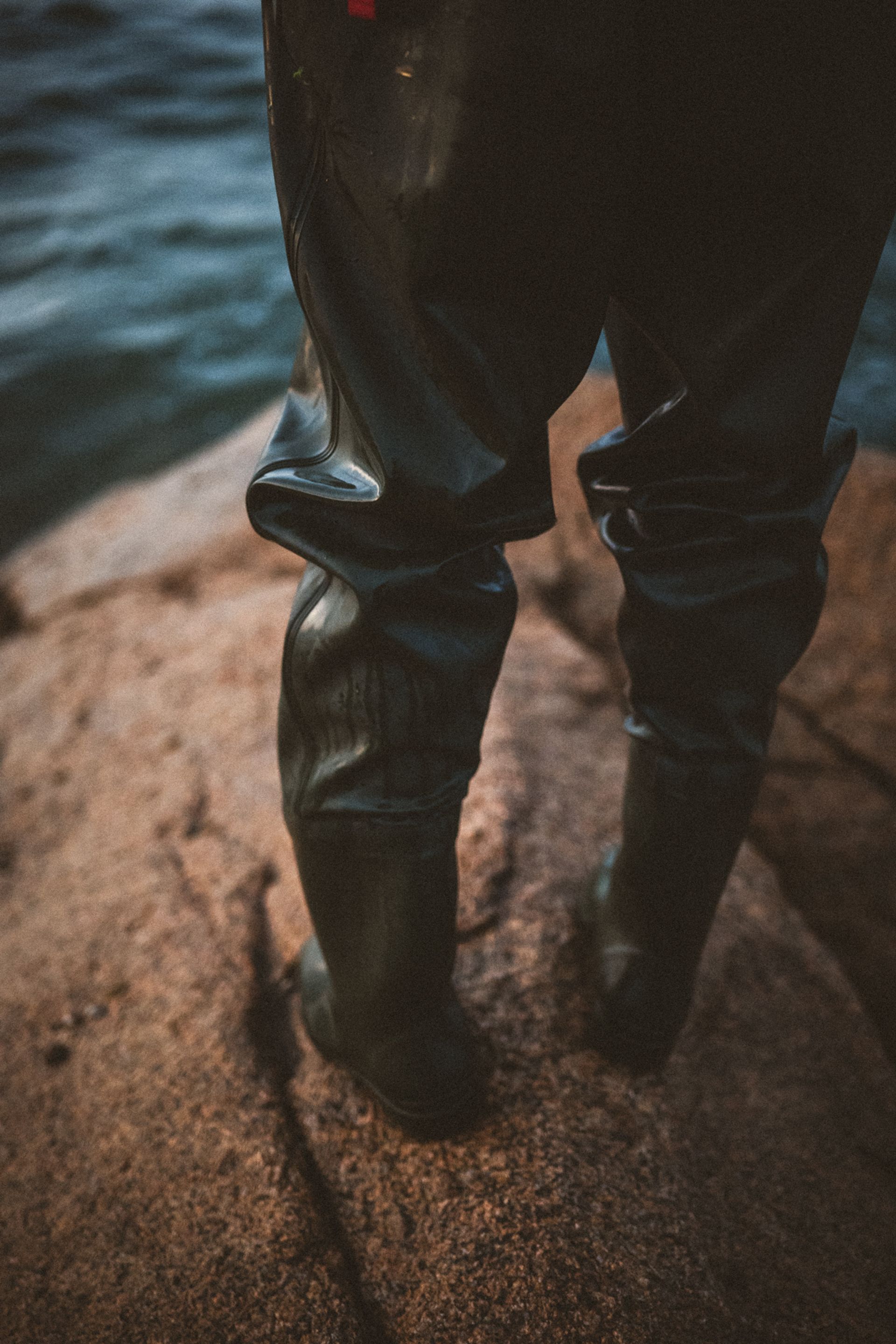 A person stands on rocky terrain wearing black waterproof waders, with water visible in the background.