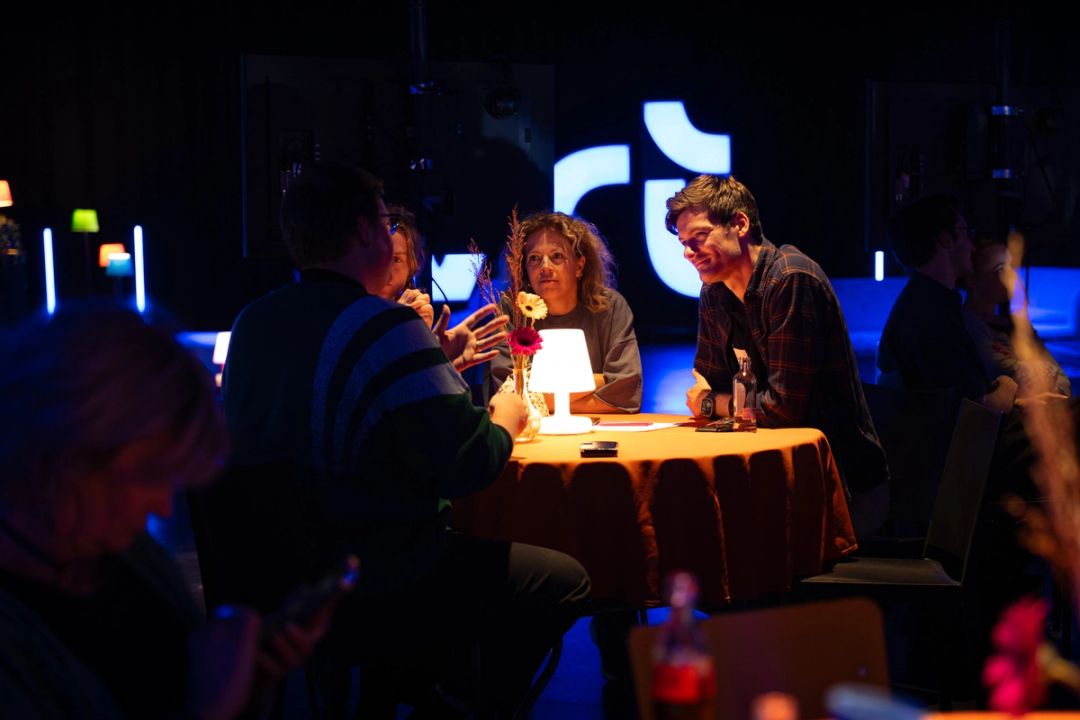 Des invites discutent a une table ronde orange avec lampe et fleurs; le logo VRT illumine brille sur la scene sombre derriere.