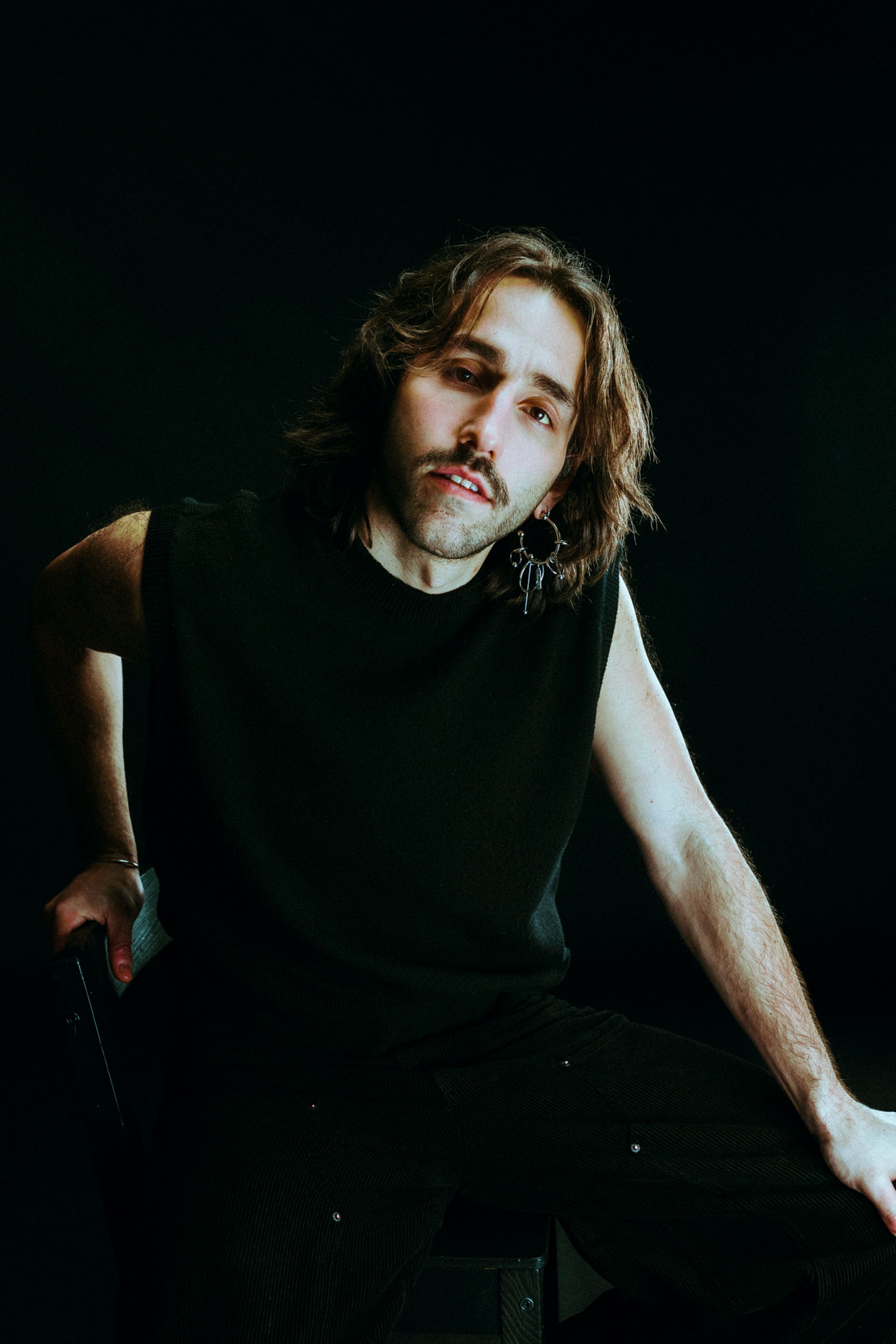 Un portrait de moi, Milo (un homme), avec des cheveux longs et un t-shirt noir, posant sur une chaise dans un fond sombre.