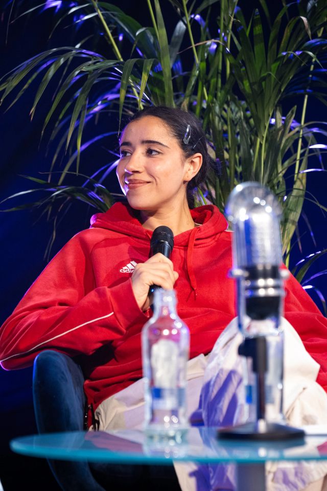 Une femme en sweat Adidas rouge sourit en tenant un micro, assise avec des micros vintage et une bouteille sur table en verre.