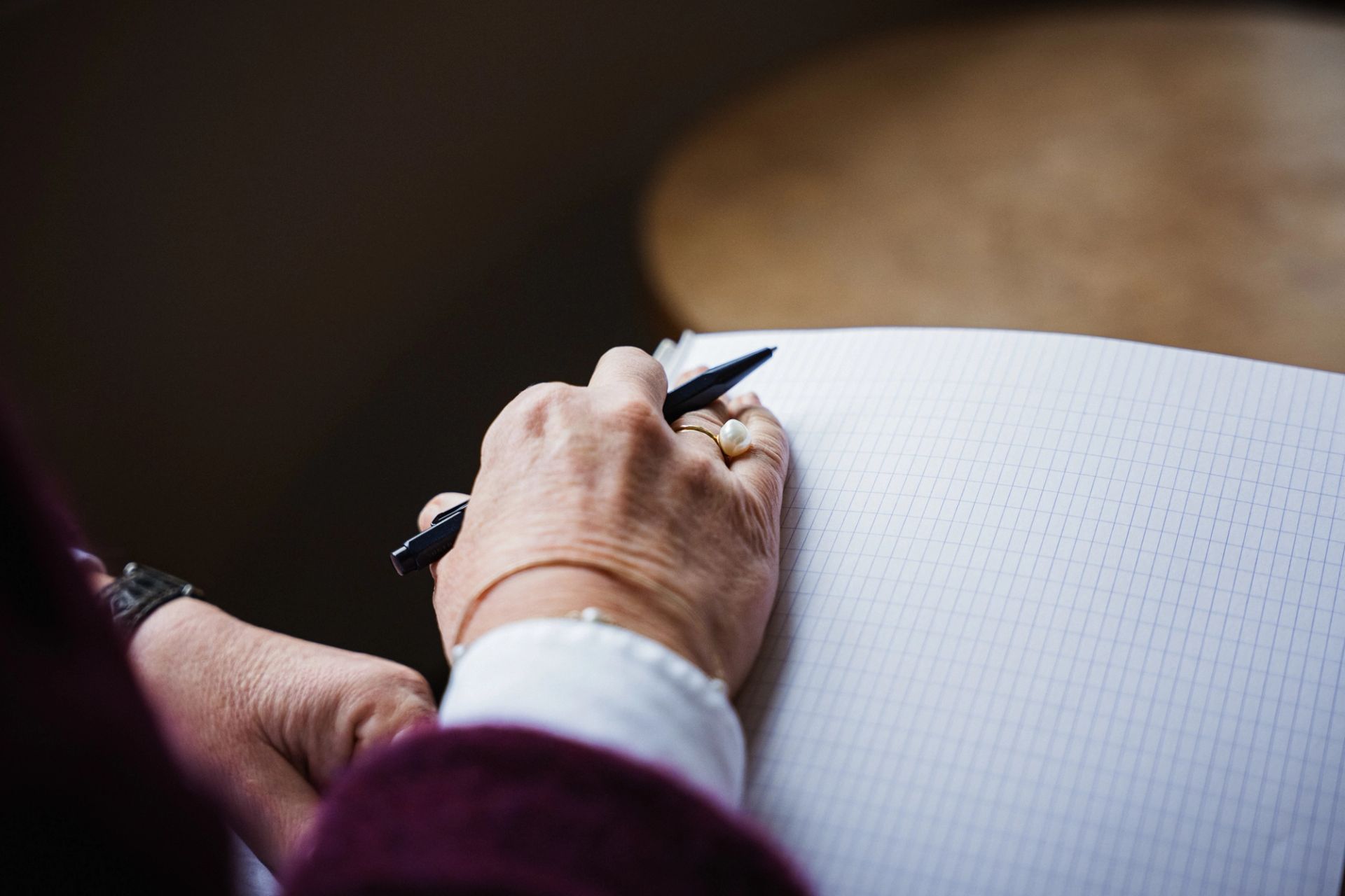 A close-up of a hand with a pearl ring writing in a grid notebook.