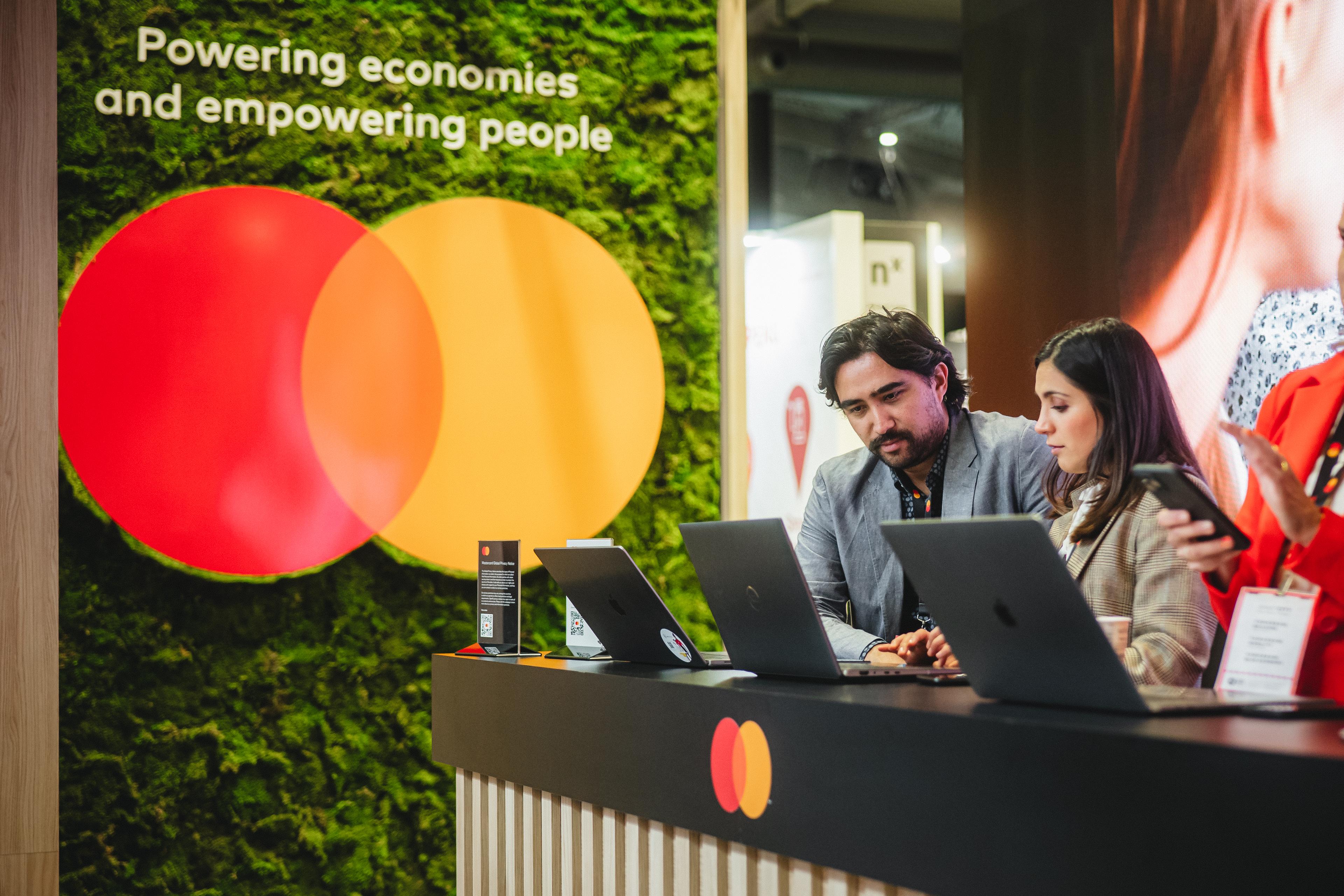 A man and a woman looking at a computer screen while standing behind the desk with the mastercard logo behind them on a green moss wall. 