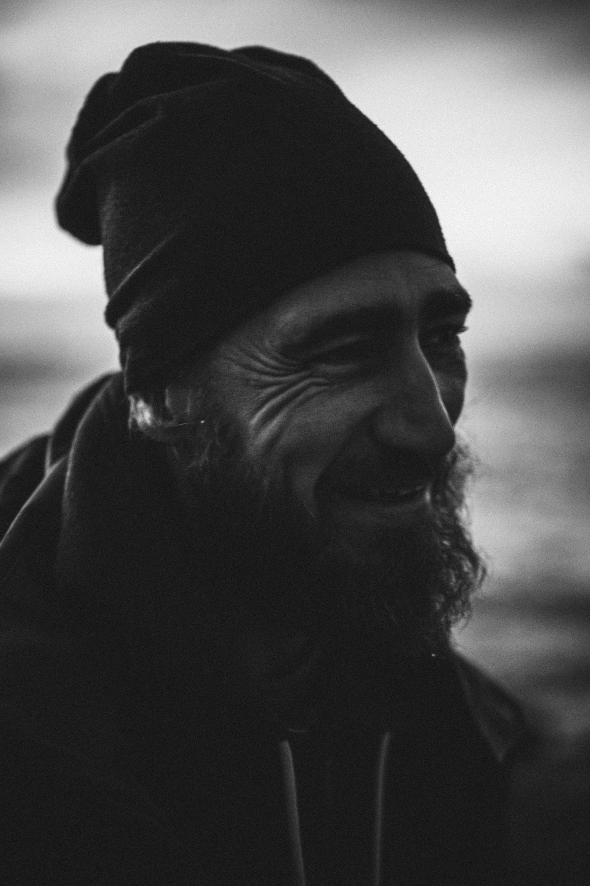A smiling man with a beard and a beanie, captured in soft focus monochrome, background blurred.