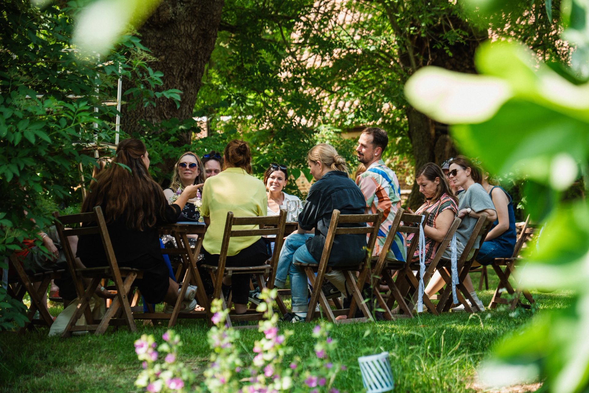 A group of colleagues sit around a long outdoor table in conversation, framed by green foliage.