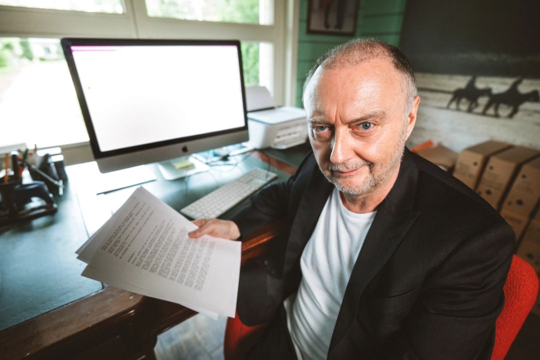Professional portrait of Luc Wyns at his desk holding documents in front of an iMac, corporate portrait photography for Knokke Off by Milo Weiler, Belgium.