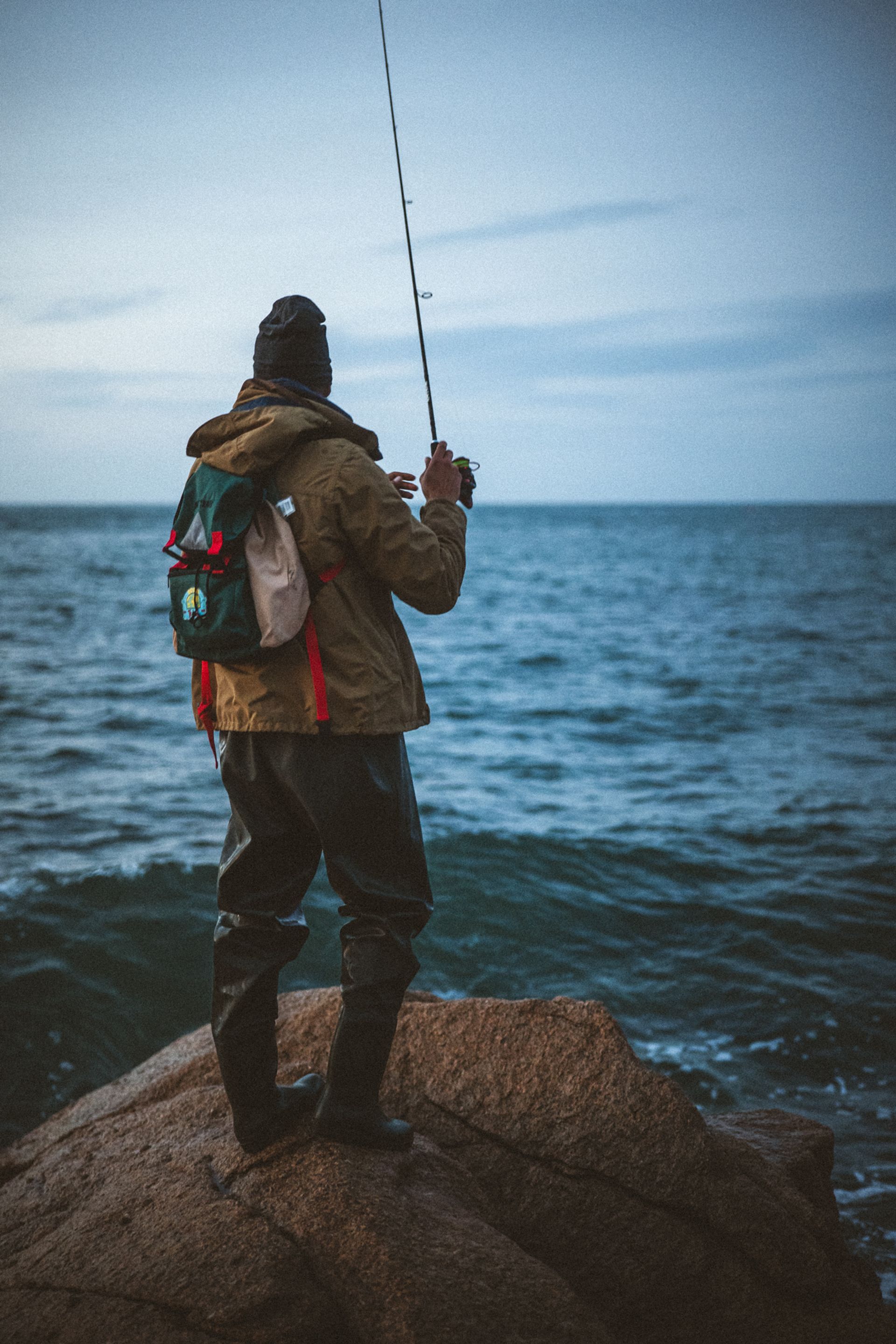 A person standing on a rock, fishing in the ocean at dusk, wearing a jacket, hat, and waders with a backpack.